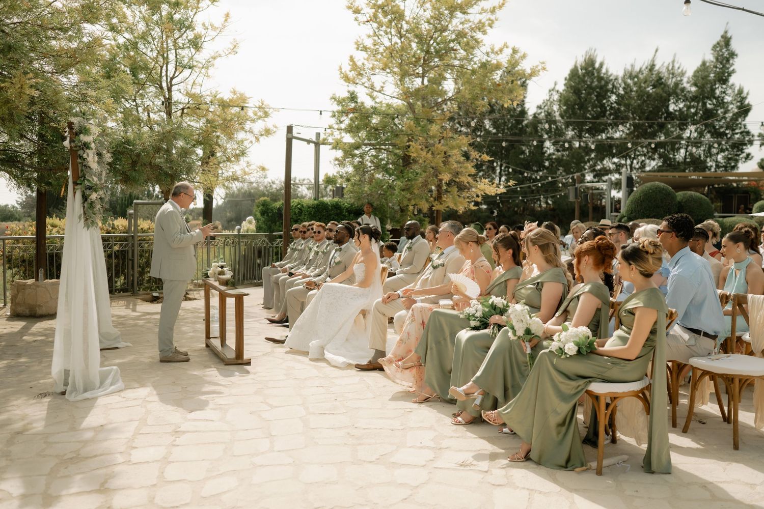 Outdoor wedding ceremony with bridesmaids in sage green dresses seated beside couple exchanging vows under sunny sky.
