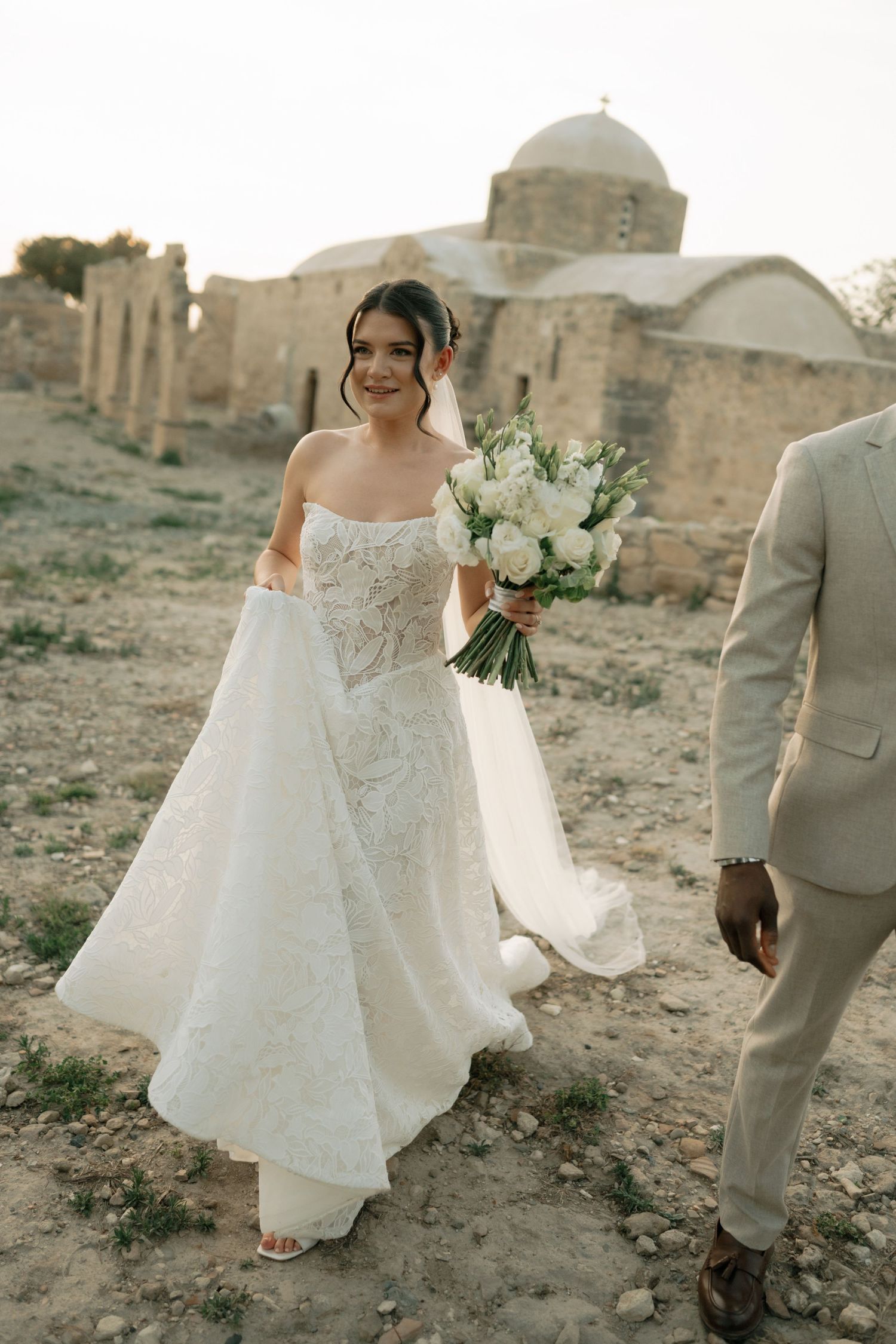 Bride in white lace gown holds bouquet near ancient stone church ruins, walking with groom in tan suit.