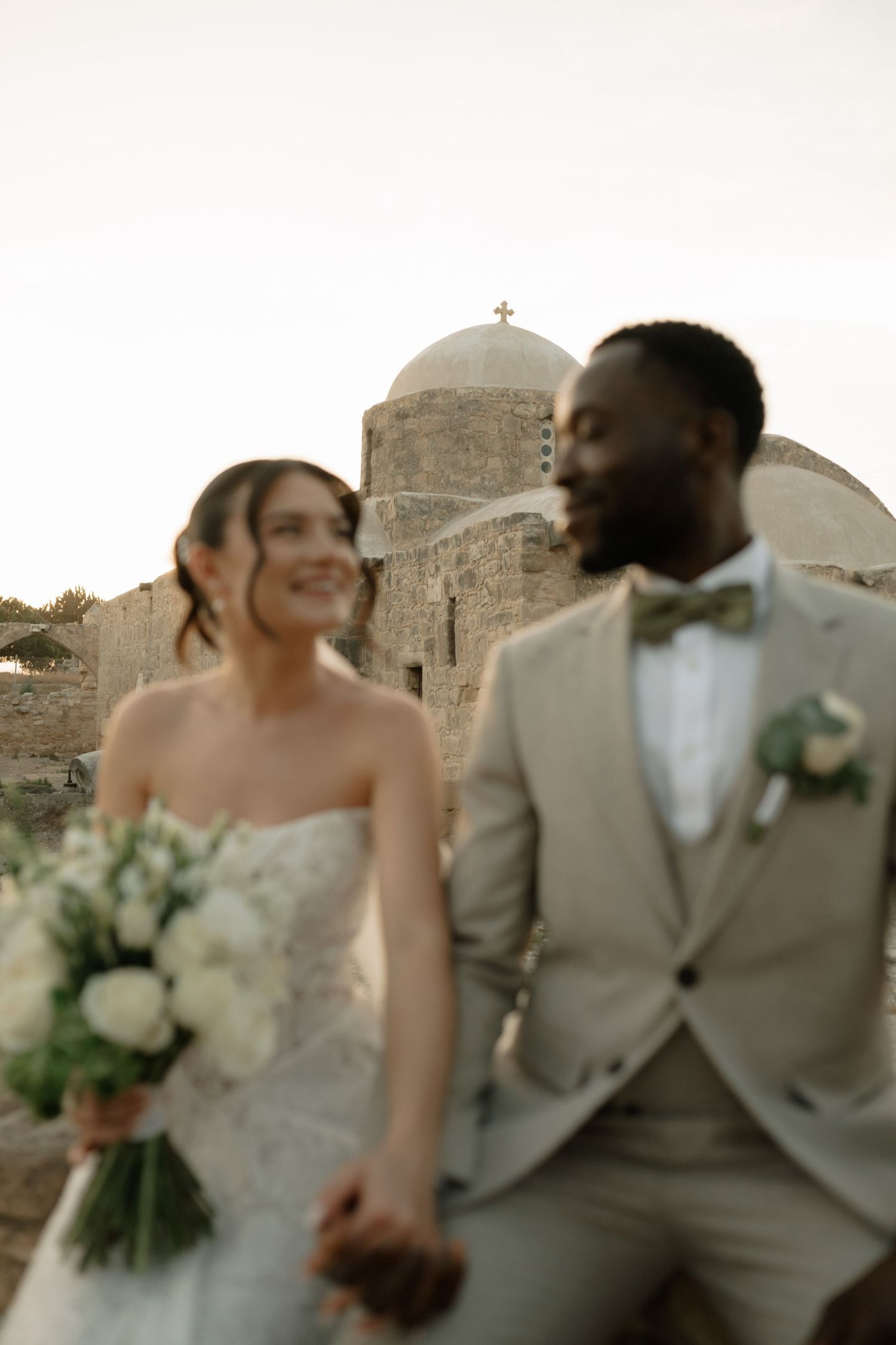 Interracial couple holding hands on wedding day, bride with white bouquet, groom in tan suit, Greek church backdrop.