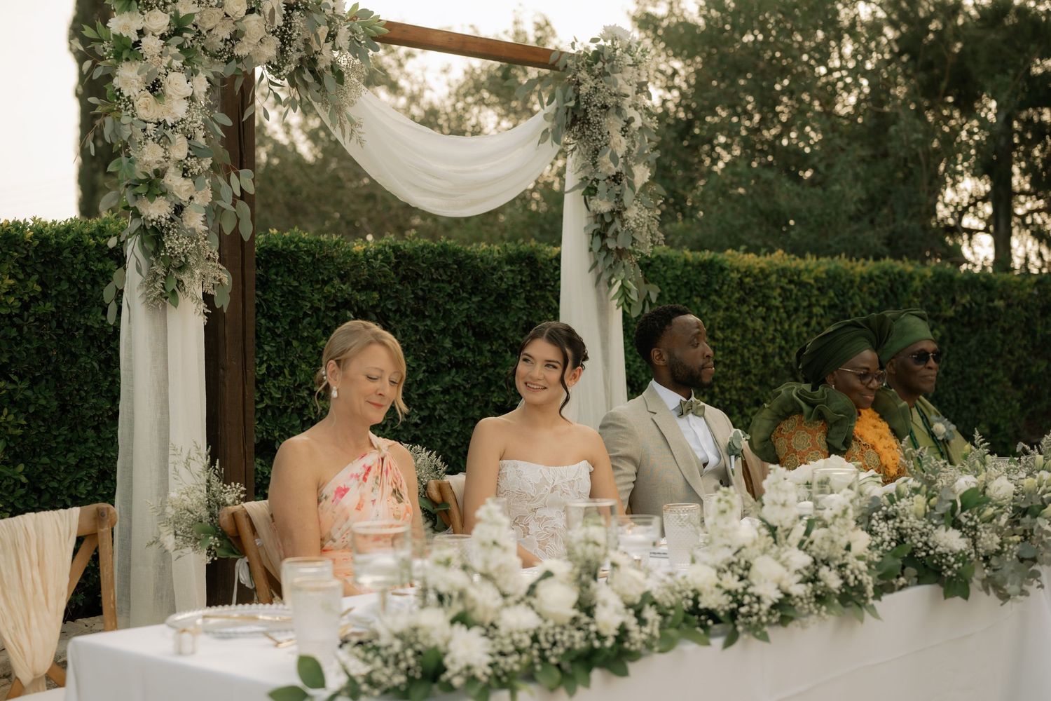Wedding party seated at a floral head table outdoors, adorned with white flowers and greenery under a draped canopy.