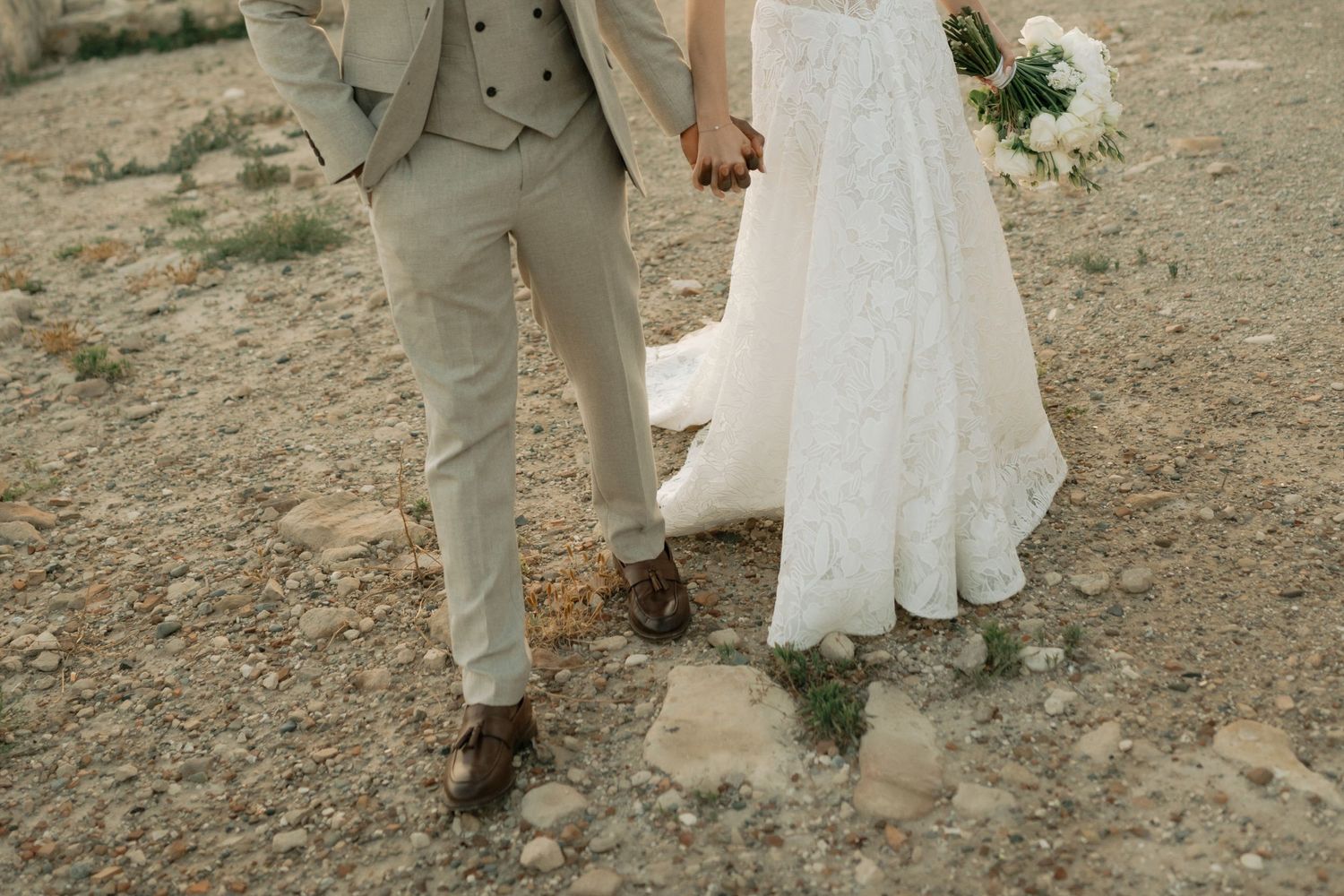 Bride in lace wedding dress and groom in beige suit holding hands on rocky terrain outdoors.