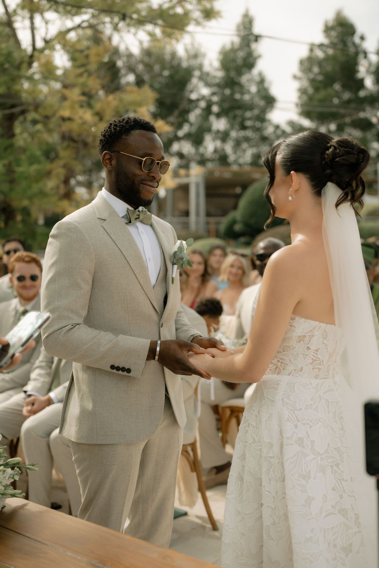 Groom in beige suit holds bride's hands during outdoor wedding ceremony with seated guests in background.