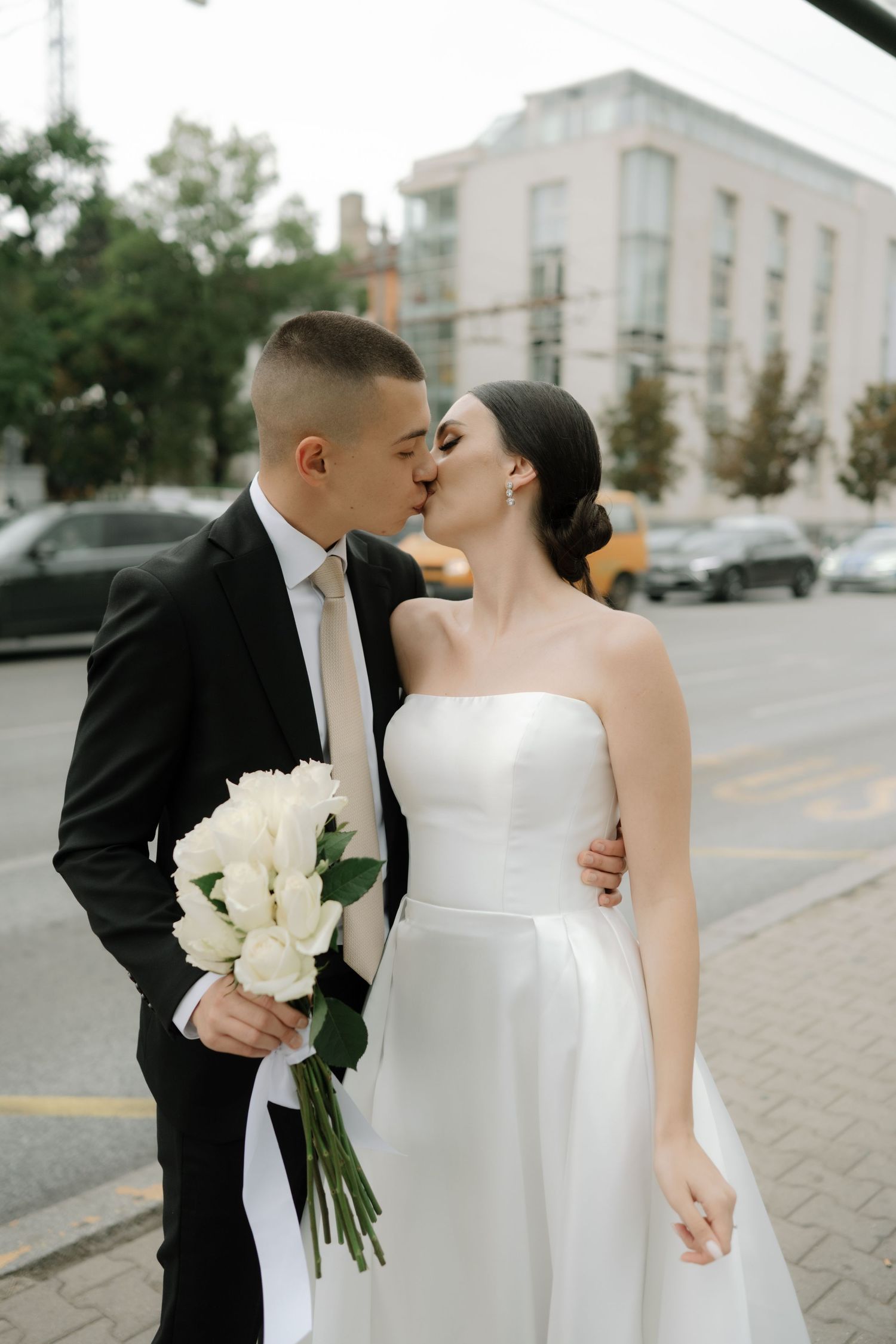 A romantic wedding couple shares intimate moments on a city street while holding white floral bouquets.