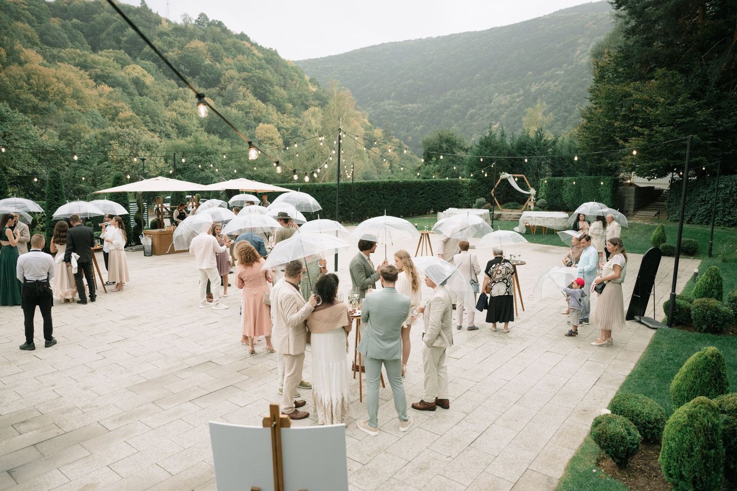 Outdoor event attendees gather with white umbrellas on a path surrounded by lush green hills on an overcast day.