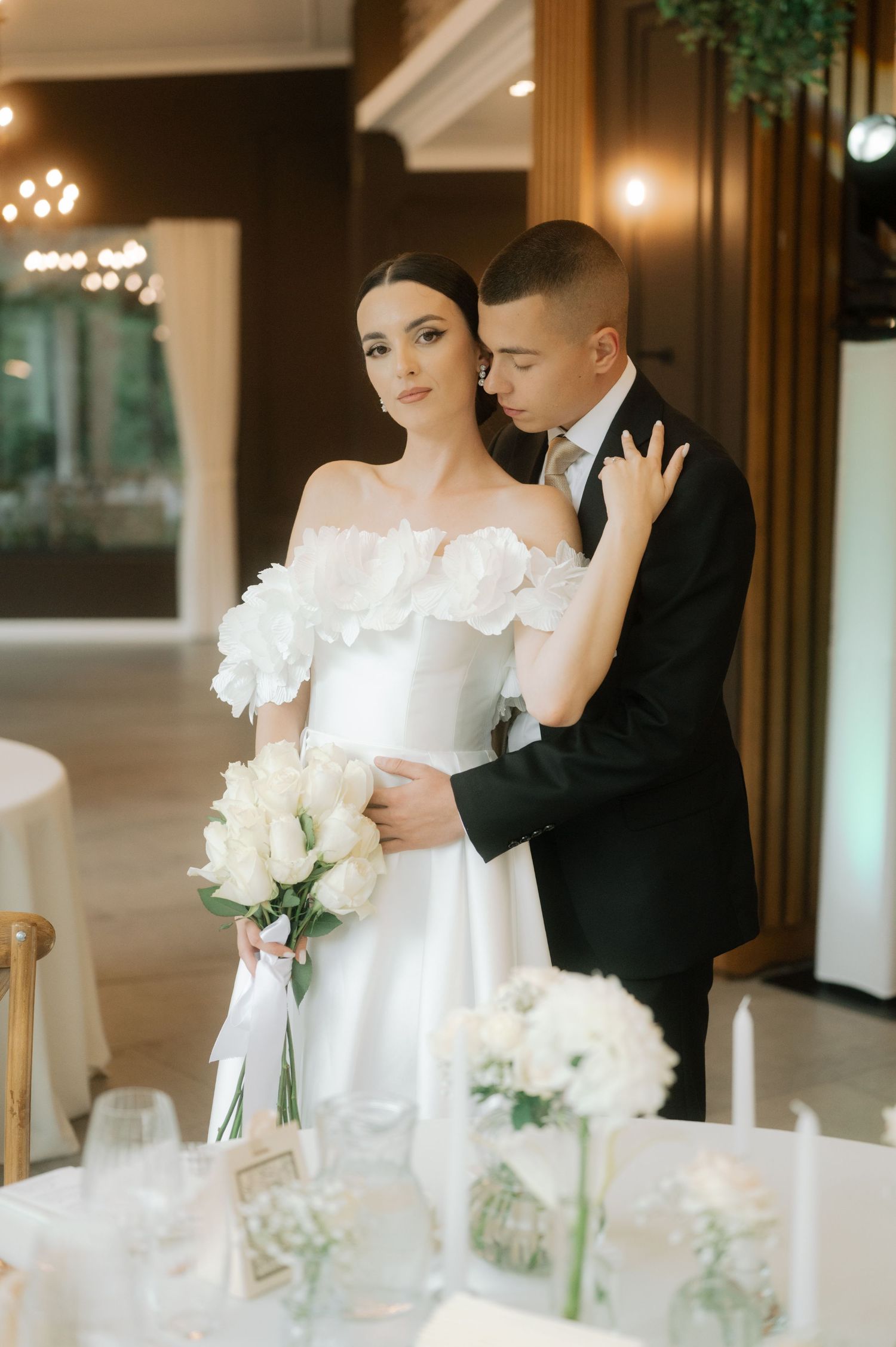 Elegant wedding couple embraces at reception venue with white floral bouquet and romantic table decor in background.