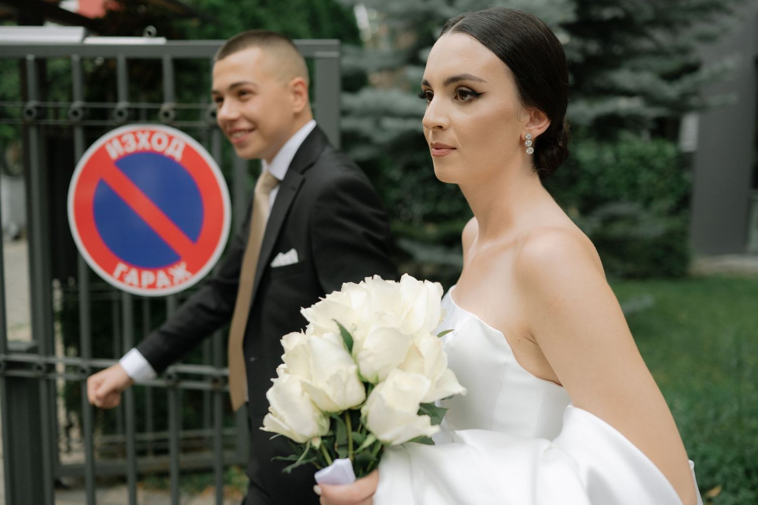 A bride and groom walk together outdoors while holding white floral bouquets near a parking sign.