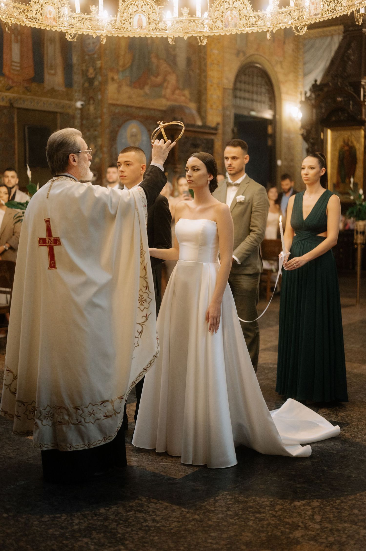 A priest performs a traditional Orthodox Christian wedding ceremony in an ornate church with a chandelier overhead.