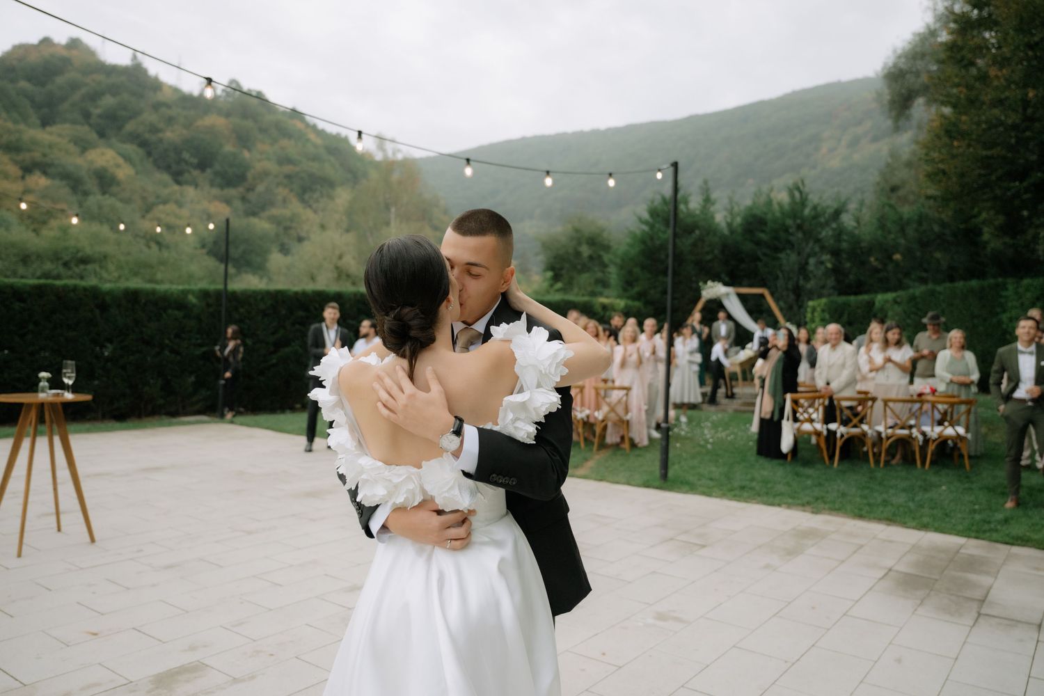 A couple shares their first dance at an outdoor mountain wedding reception with string lights and guests watching.