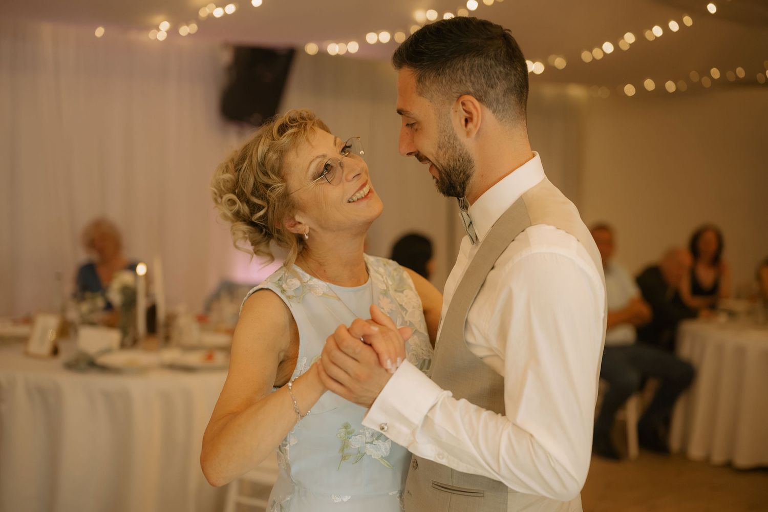 Couple sharing their first dance at a wedding reception with fairy lights and guests in the background.