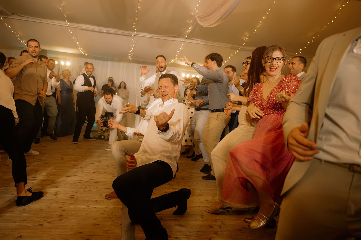 Guests dancing energetically at a wedding reception in a decorated tent with string lights.