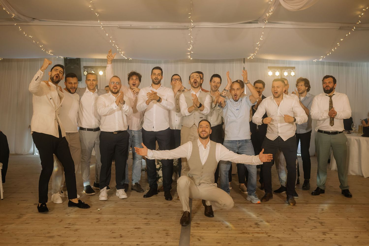 Group of groomsmen posing together at a wedding reception, smiling and celebrating with fairy lights in background.