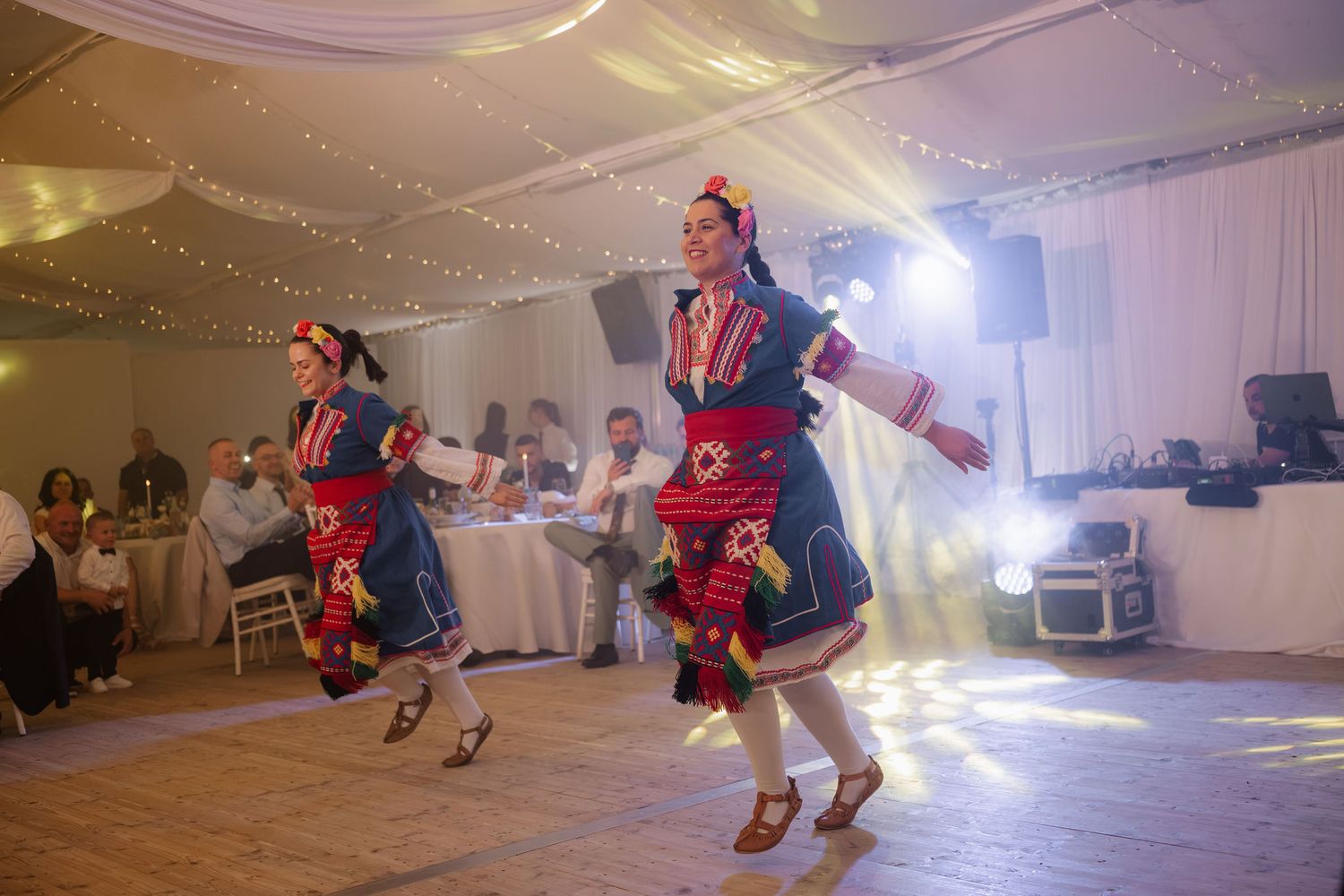 Two women in colorful traditional folk costumes perform an energetic cultural dance at an elegant wedding reception.