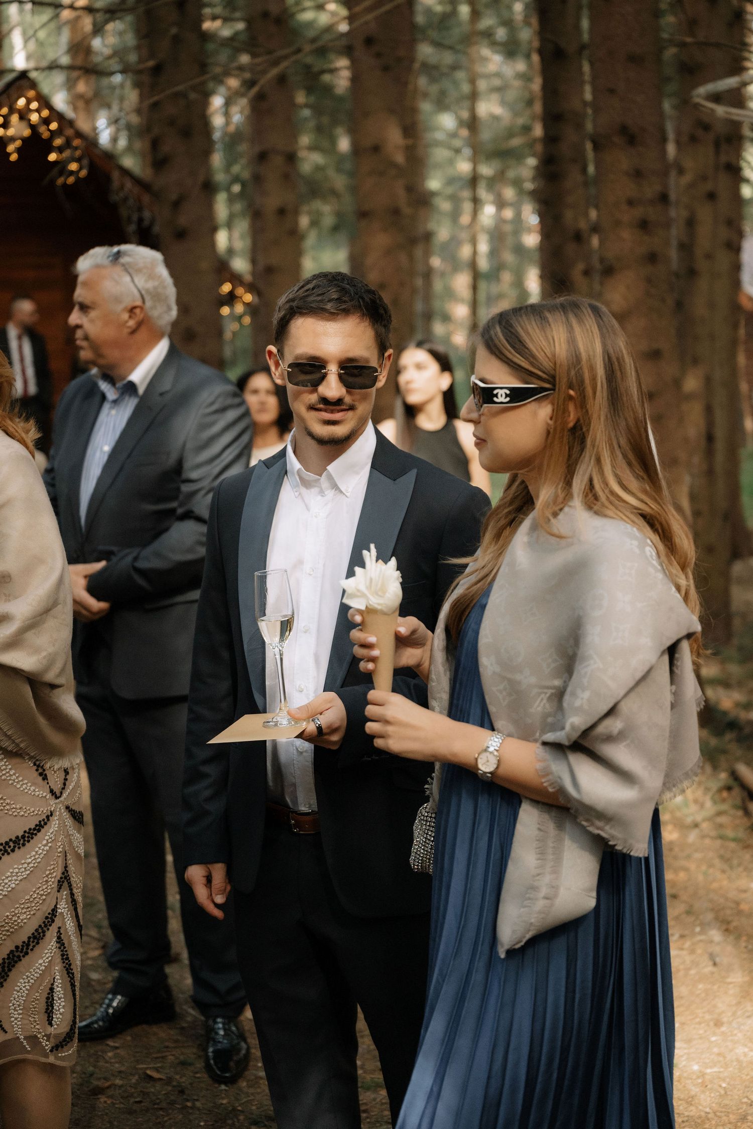 Elegantly dressed guests at an outdoor forest wedding, man in suit with sunglasses holding champagne flute.