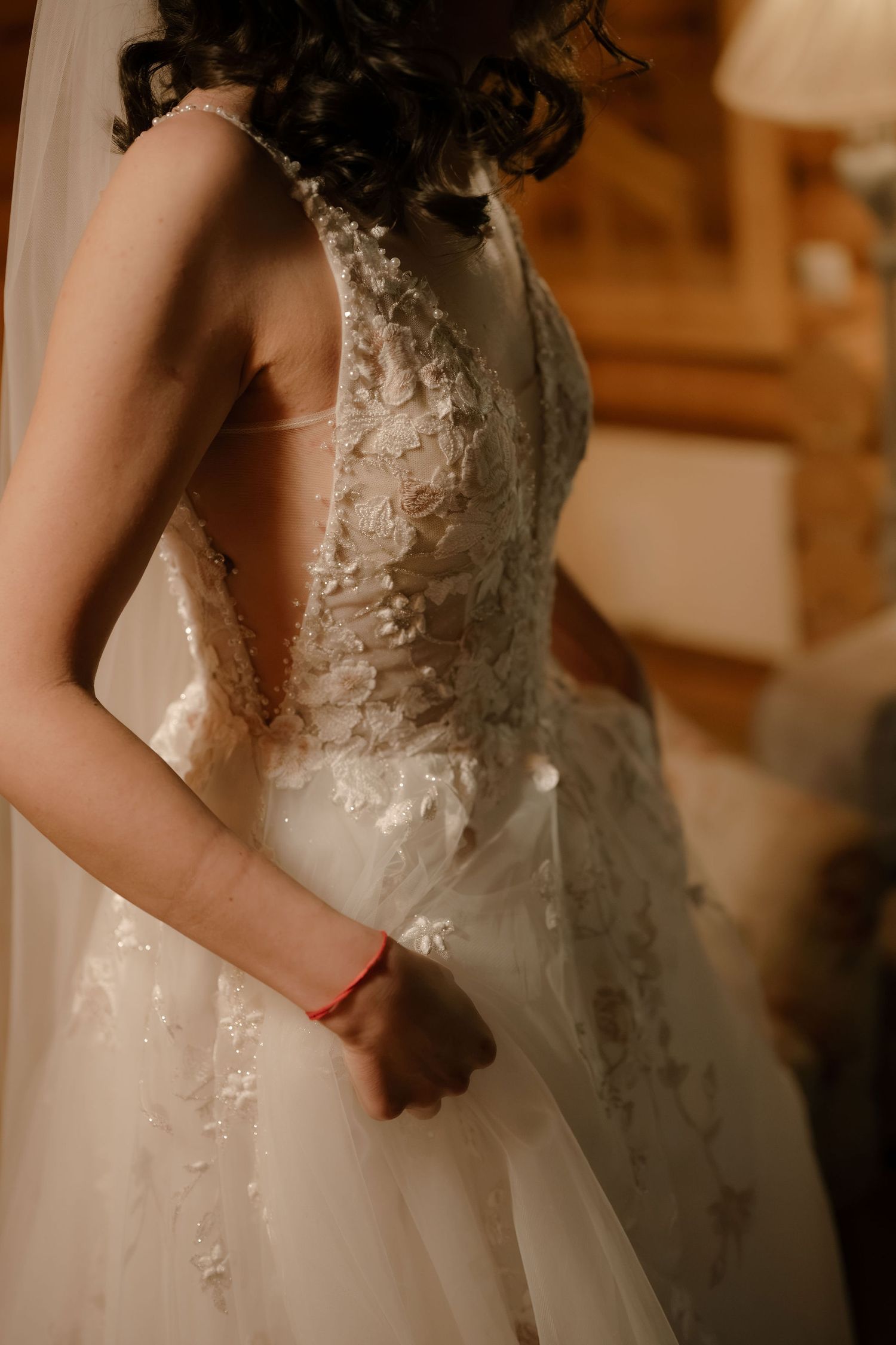 Bride in elegant lace wedding dress with floral appliques and tulle skirt, viewed from behind in warm indoor light.