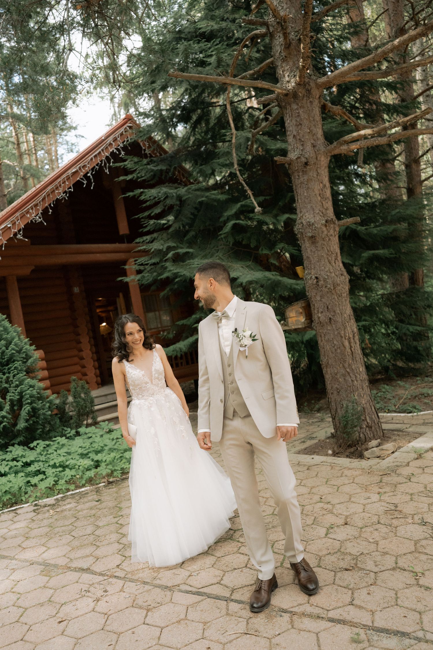 A bride in a white tulle gown and groom in a beige suit hold hands while strolling through a forest wedding venue.