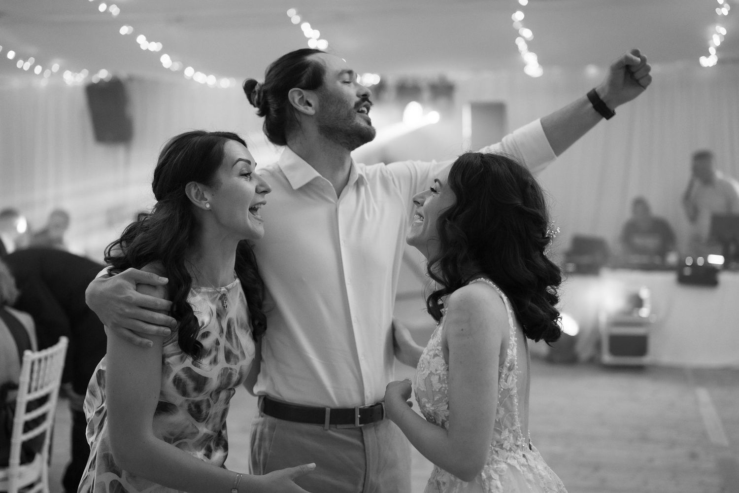 Black and white photo of a groom taking a selfie with two women laughing at a wedding reception.