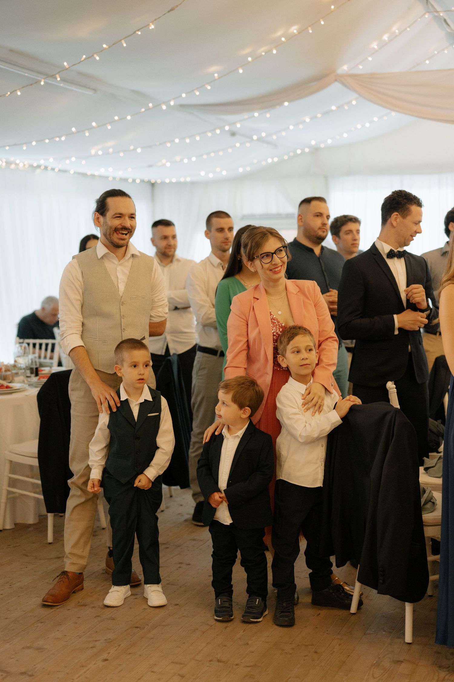 Family with two young boys smiling at a wedding reception in a decorated tent venue with string lights.