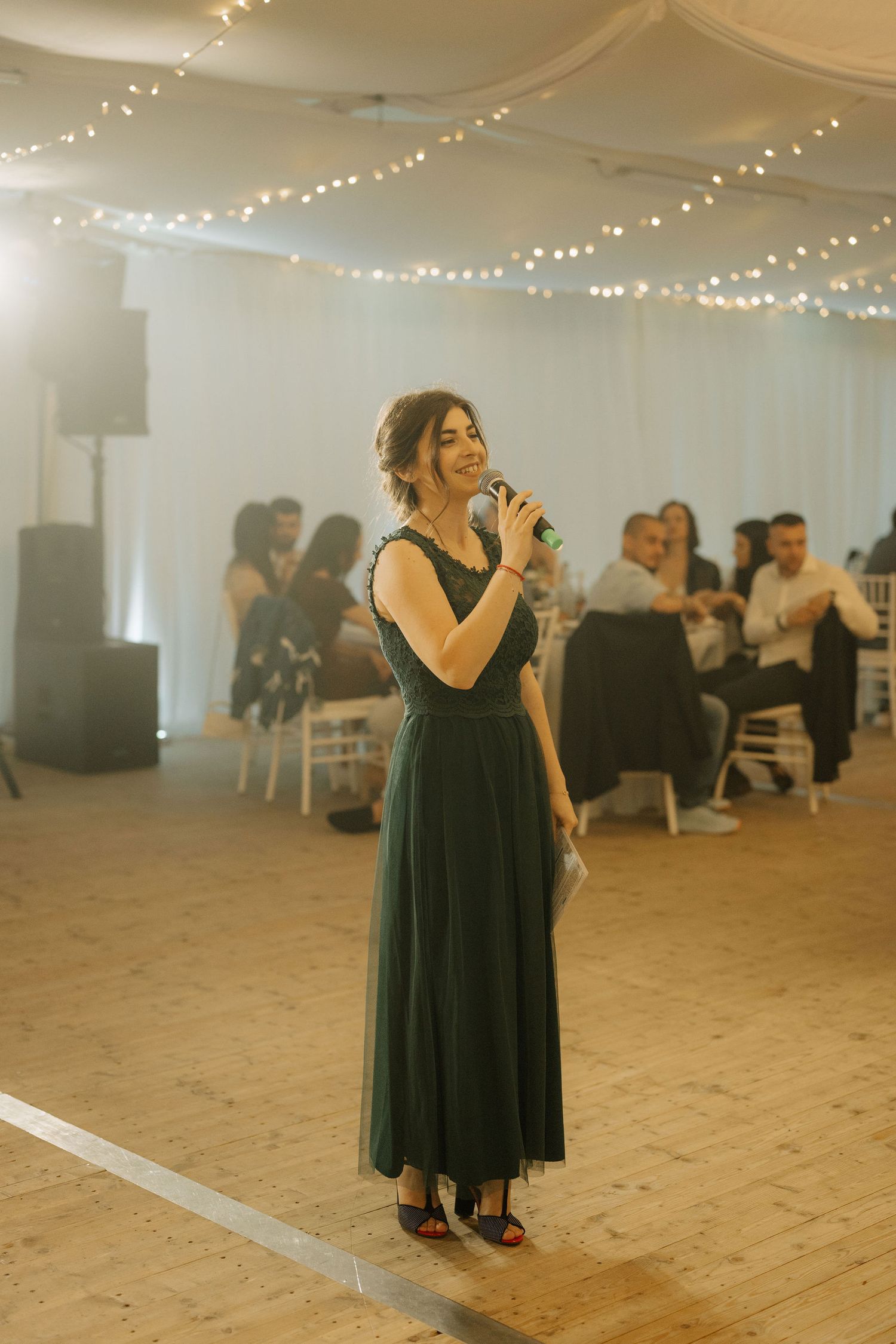 Woman in green dress speaking into microphone at elegant wedding reception with fairy lights.