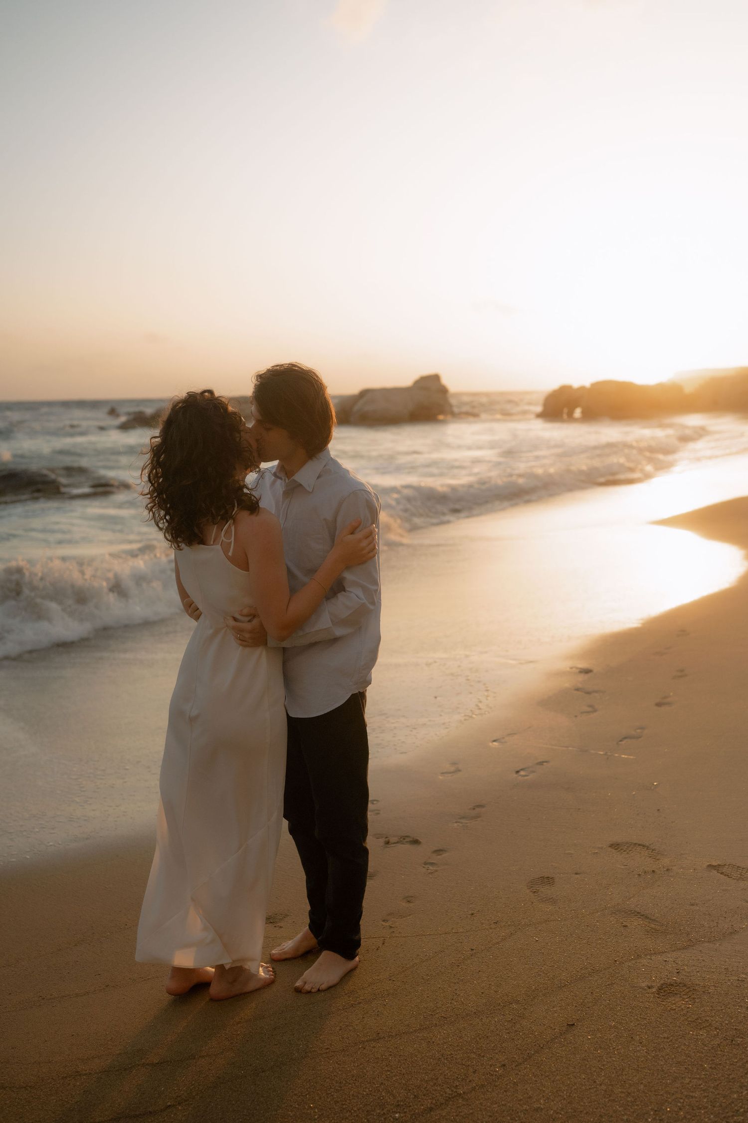Couple embracing and kissing on a sandy beach at golden sunset with ocean waves in the background.