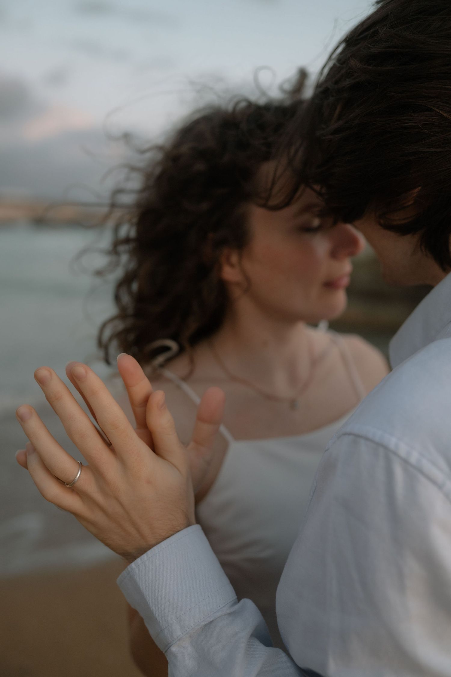 Couple embracing on beach at dusk, woman wearing ring, curly hair blowing in wind, romantic seaside engagement photo.