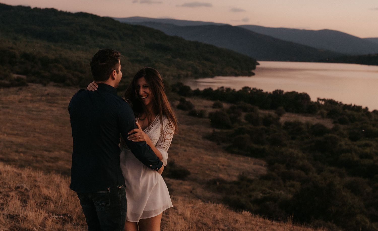 Two figures share an intimate moment against a dusky mountain landscape with a lake in the background.