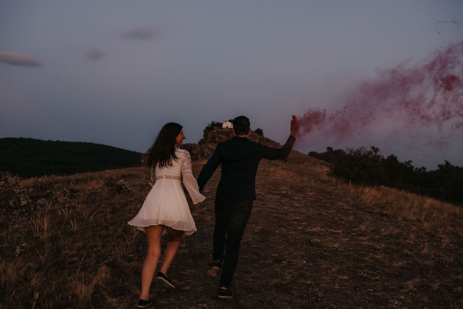Two people walk along a hilltop path at sunset with dramatic red smoke effects.