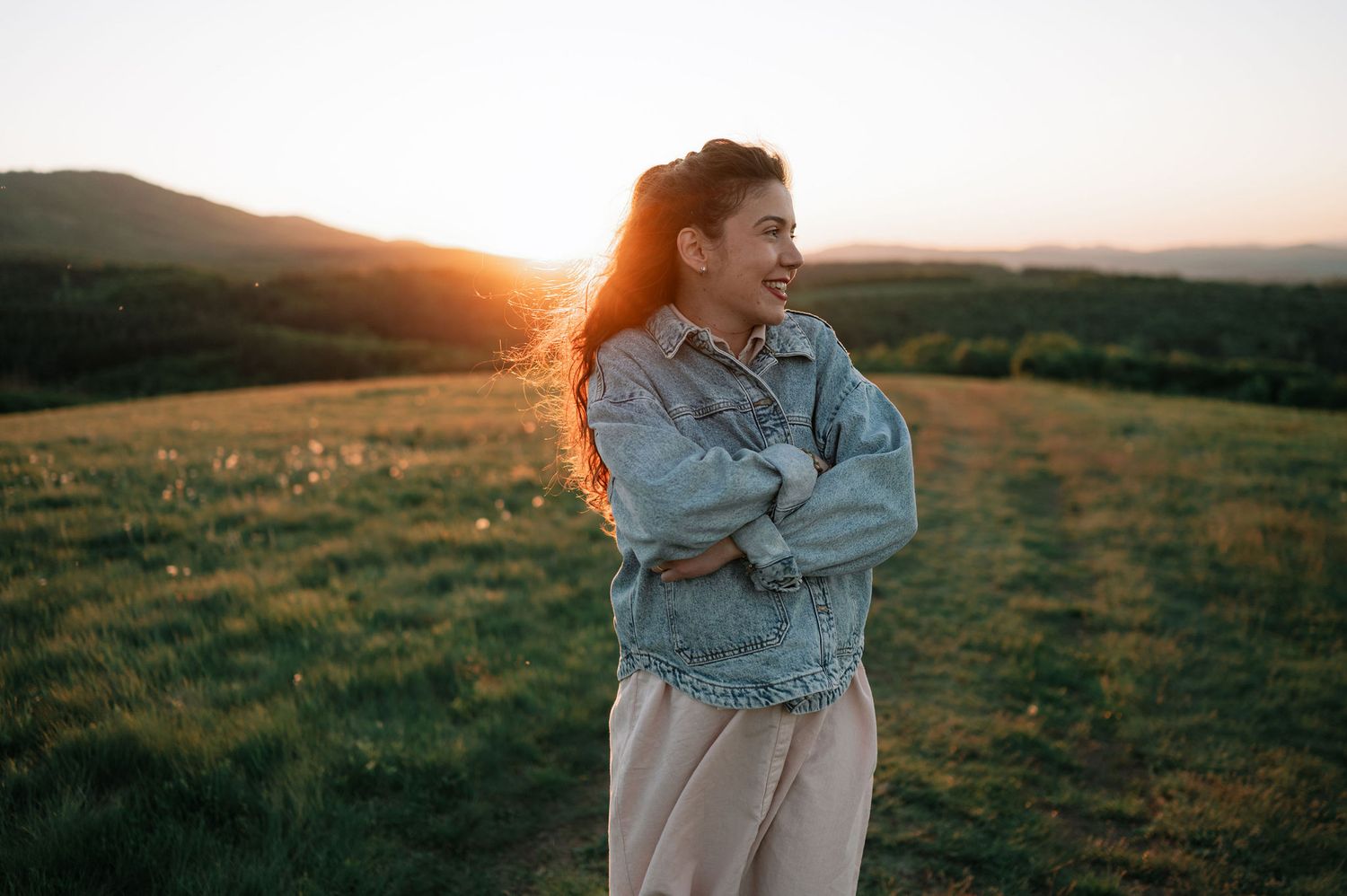 A person wearing denim embraces themselves while standing in a golden sunlit field at sunset.