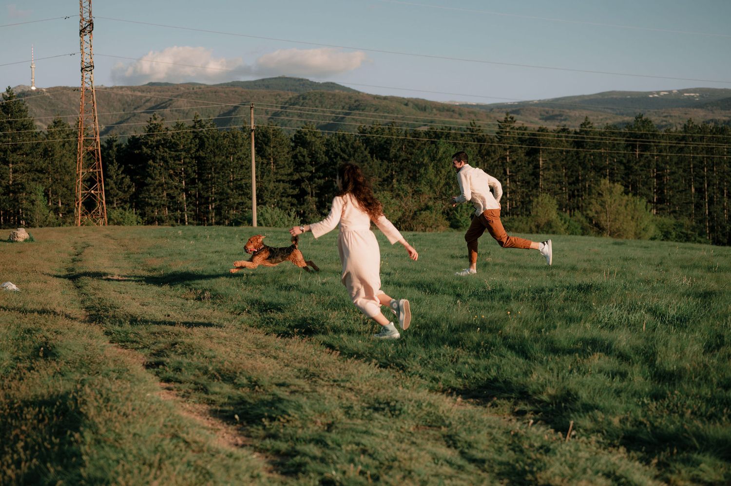 People running and playing in a grassy field with mountains and forest in the background on a sunny day.