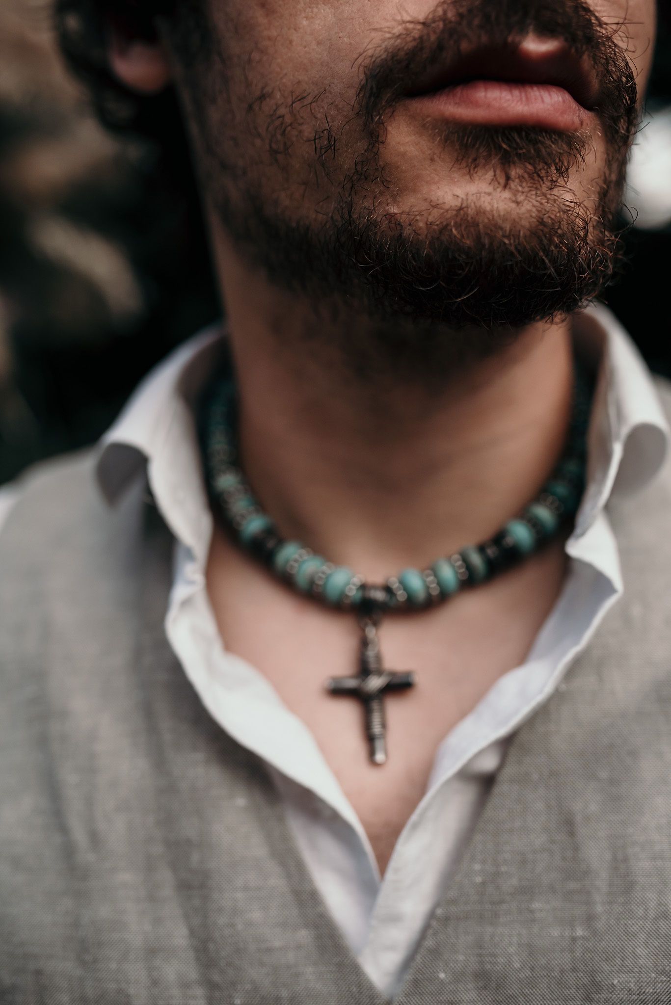 Detail of a beaded turquoise choker necklace with cross pendant against white collar and gray vest.