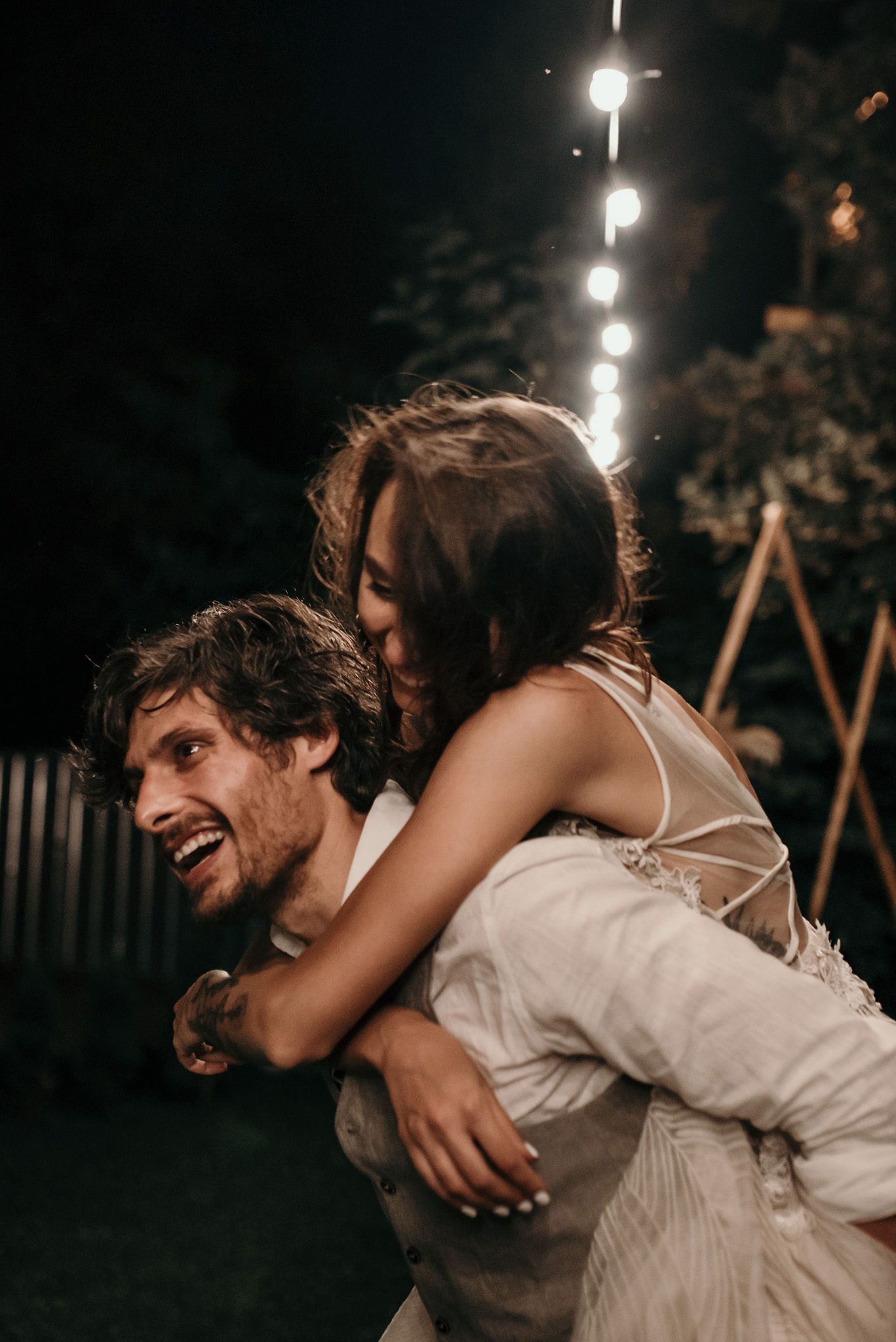 A couple shares a joyful moment at night with string lights glowing in the background during a piggyback ride.