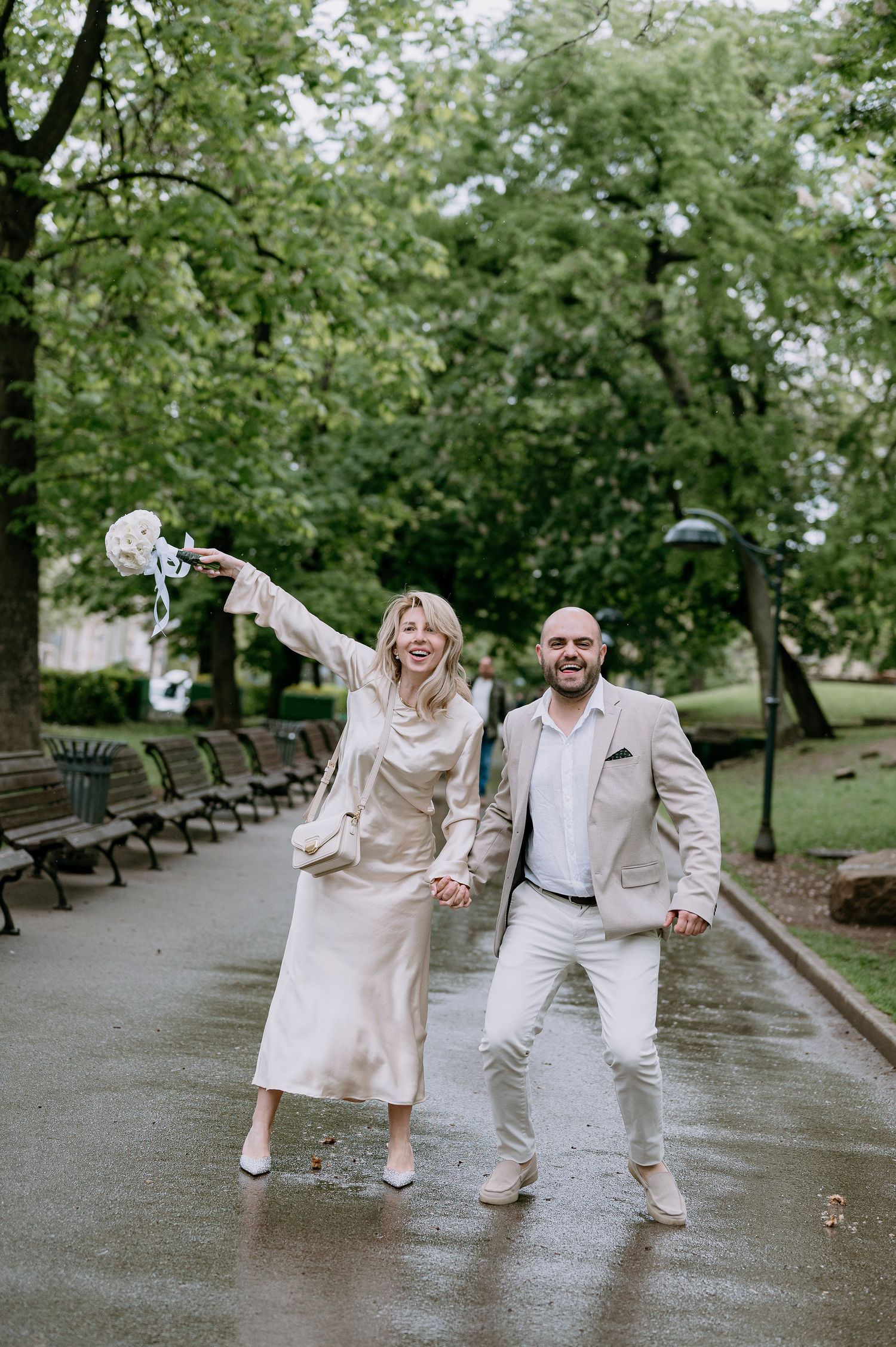 A playful couple in coordinated white outfits dances and poses on a wet park pathway.