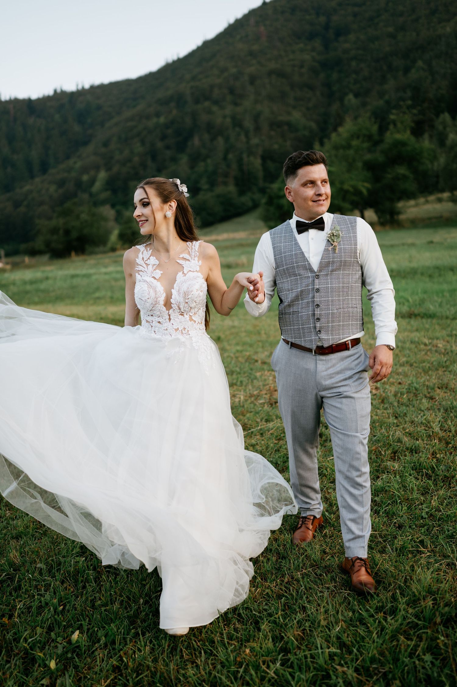 Bride in flowing lace gown and groom in vest walk joyfully across mountain meadow.