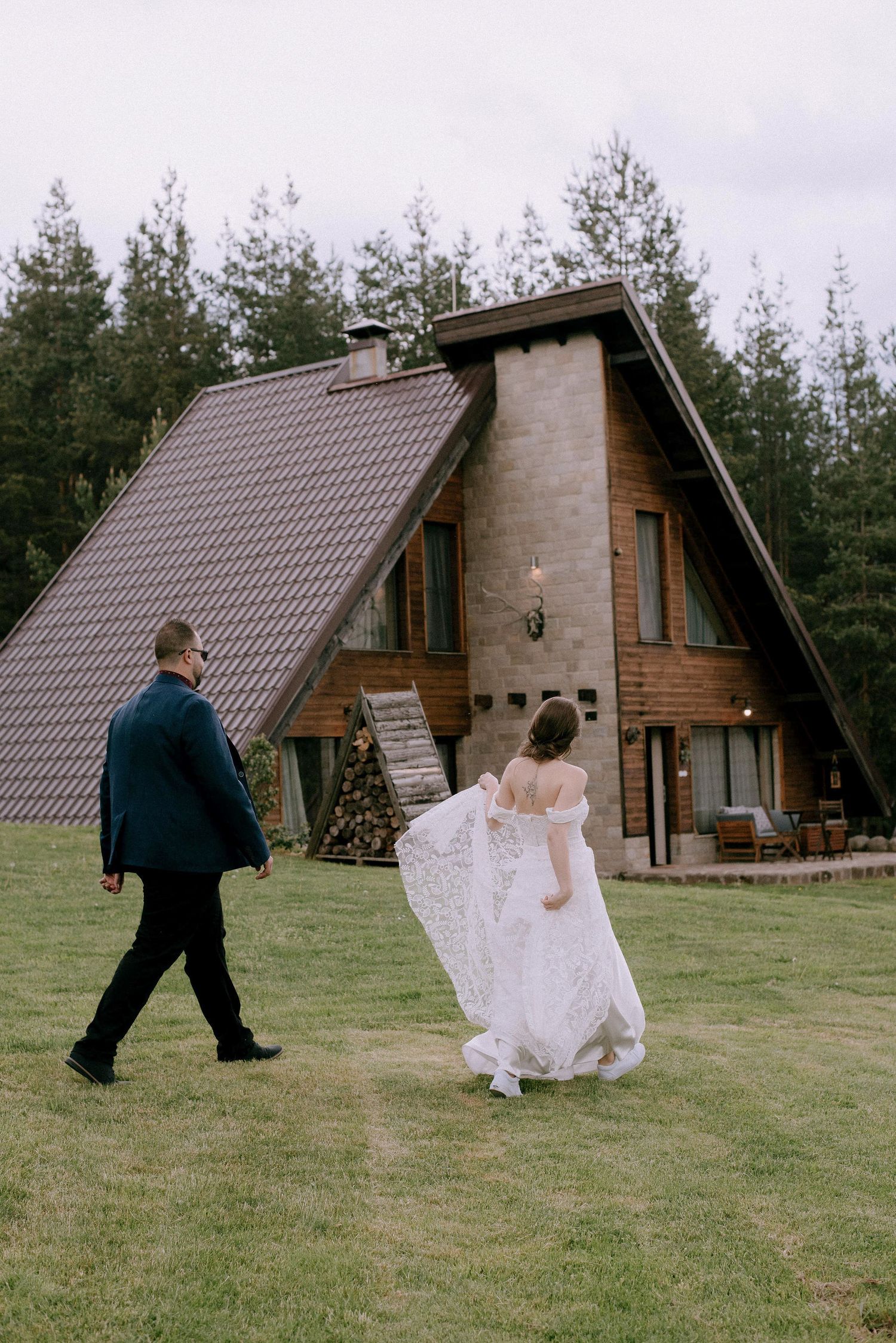 A couple walks across a grassy lawn towards a rustic A-frame cabin in the woods.