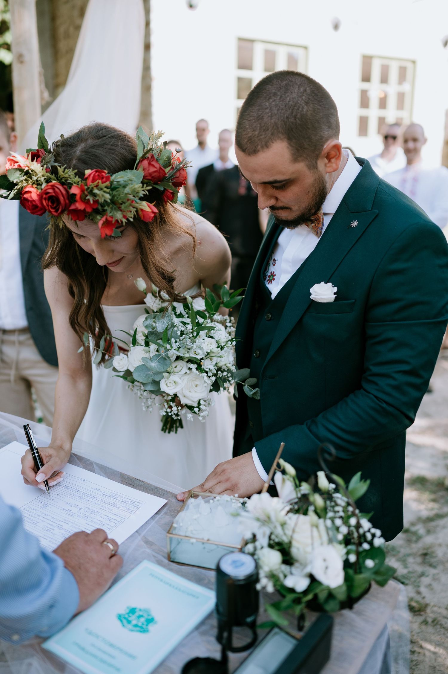 Couple signing marriage documents at an outdoor ceremony with white and red floral arrangements.