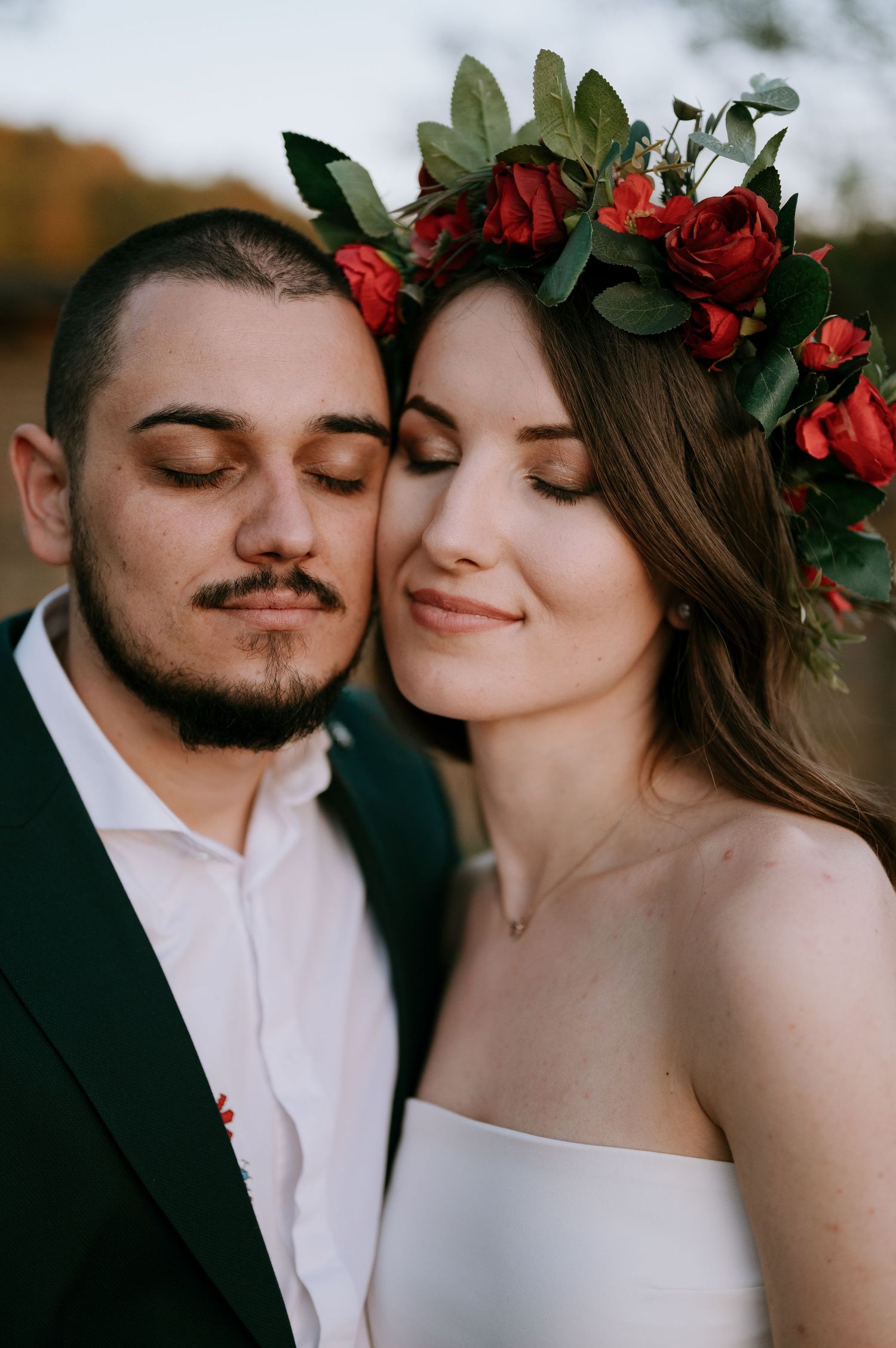 Romantic close-up of newlyweds with red floral crown against natural backdrop.