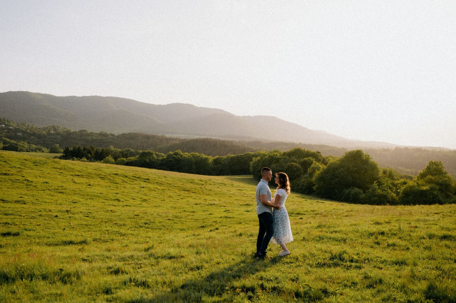 A panoramic view shows two people in a vast green meadow at sunset.