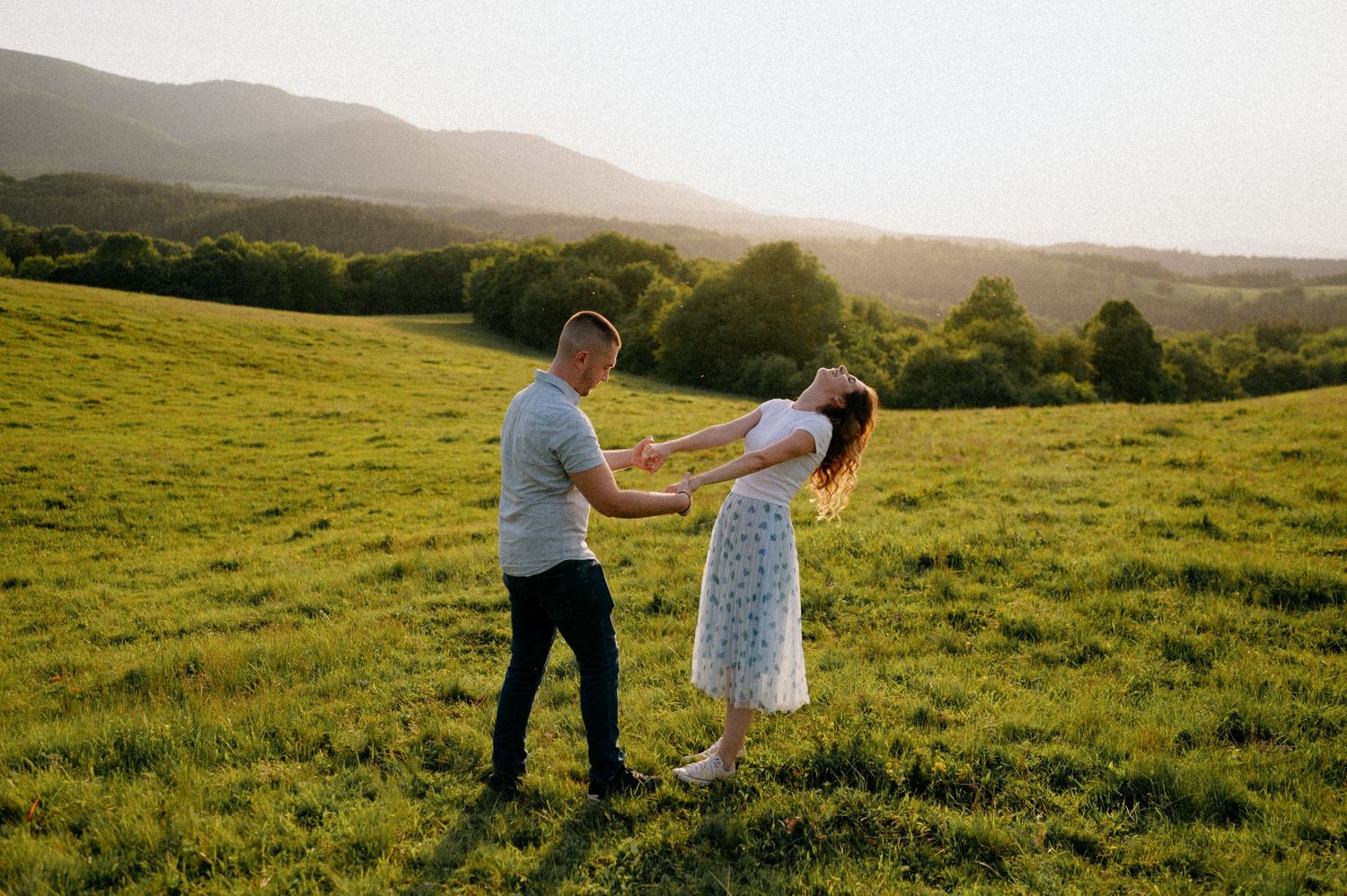 Couple dancing together in a sunny green meadow with rolling hills in the background.