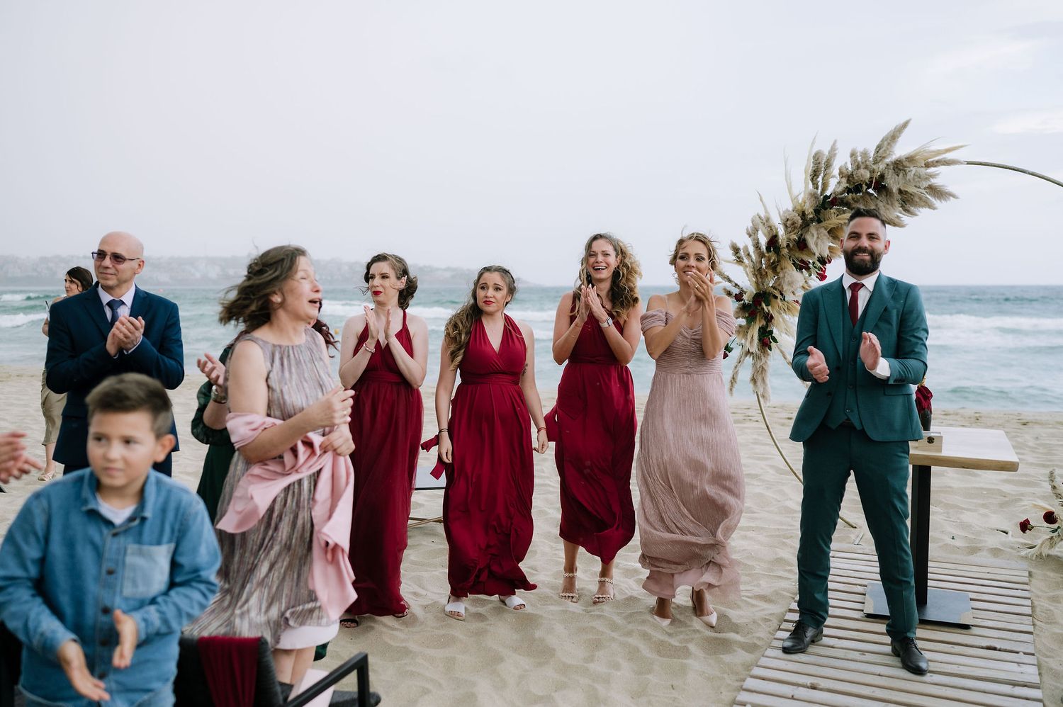 Wedding party members in burgundy and teal attire gather on beach boardwalk for photos.