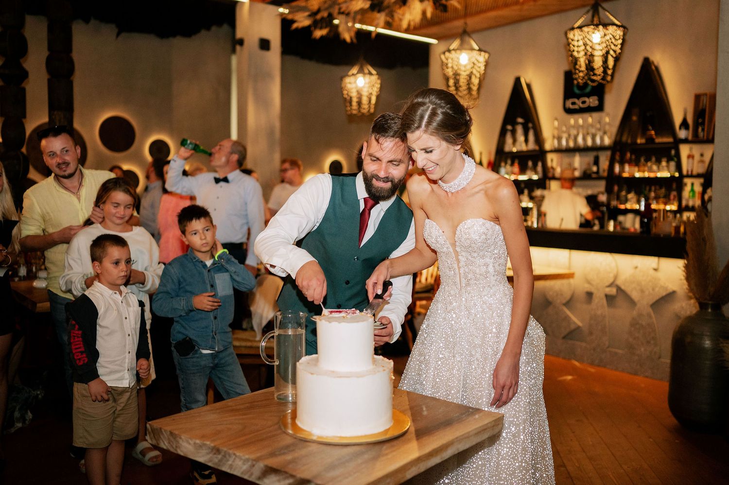 A couple cuts their elegant white wedding cake together at a reception with vintage lighting.