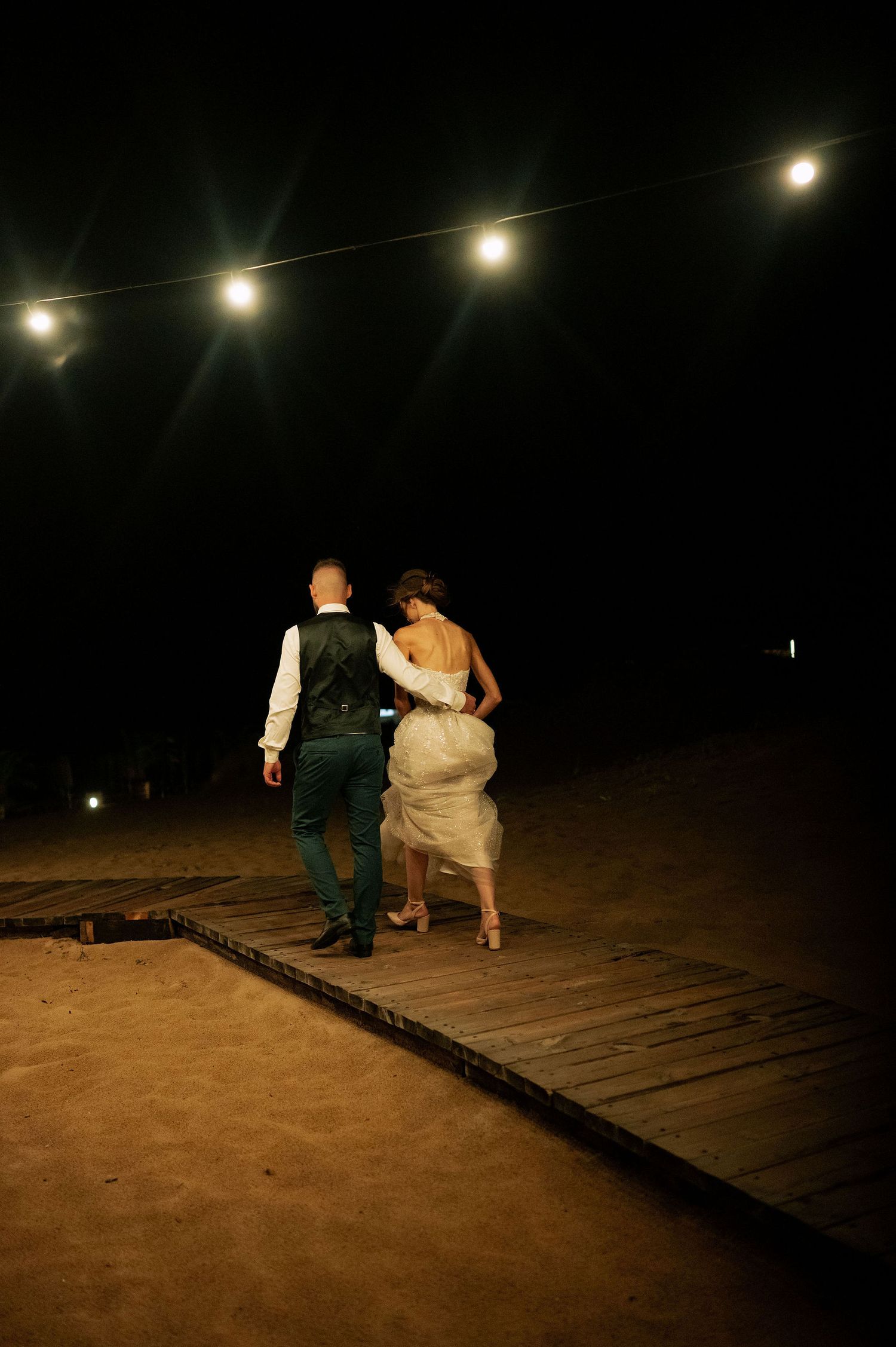 Couple walking along a wooden boardwalk under string lights at night.