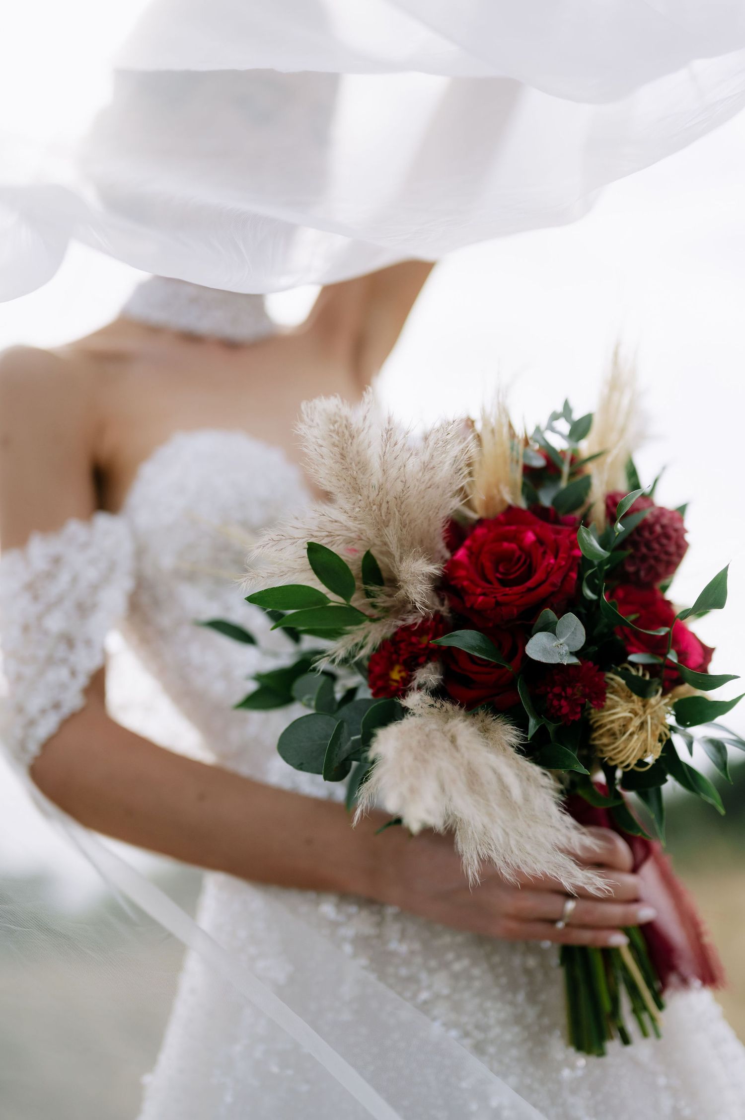 Close-up of a bridal bouquet featuring deep red roses, pampas grass, and greenery against a white dress.