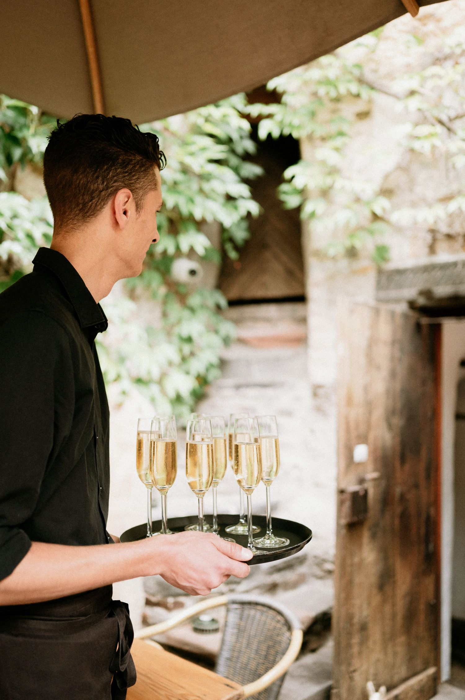 Server carries tray of champagne glasses on outdoor patio with greenery.