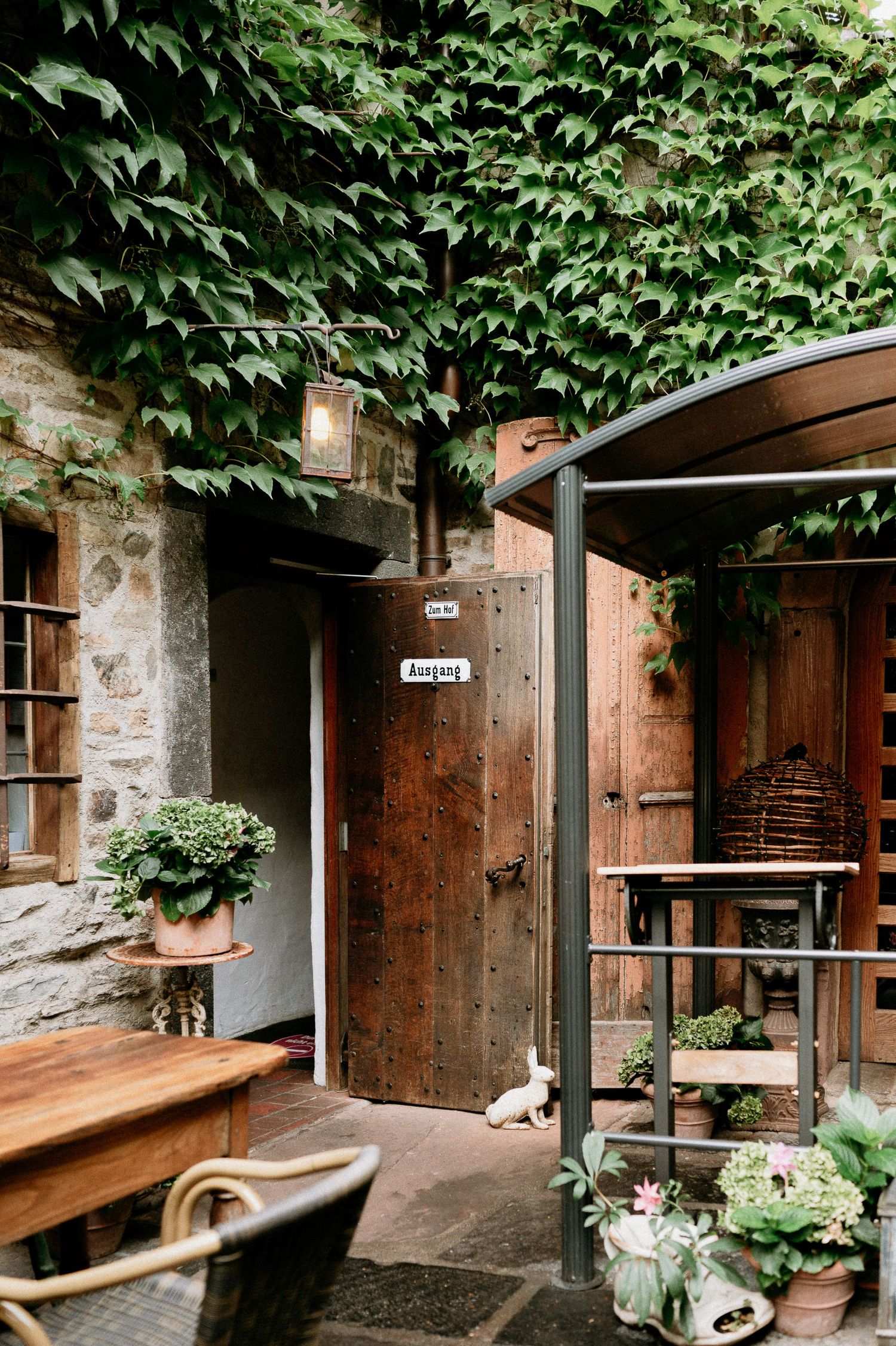 Stone and brick entryway with wooden doors and green foliage growing overhead creates a rustic ambiance.