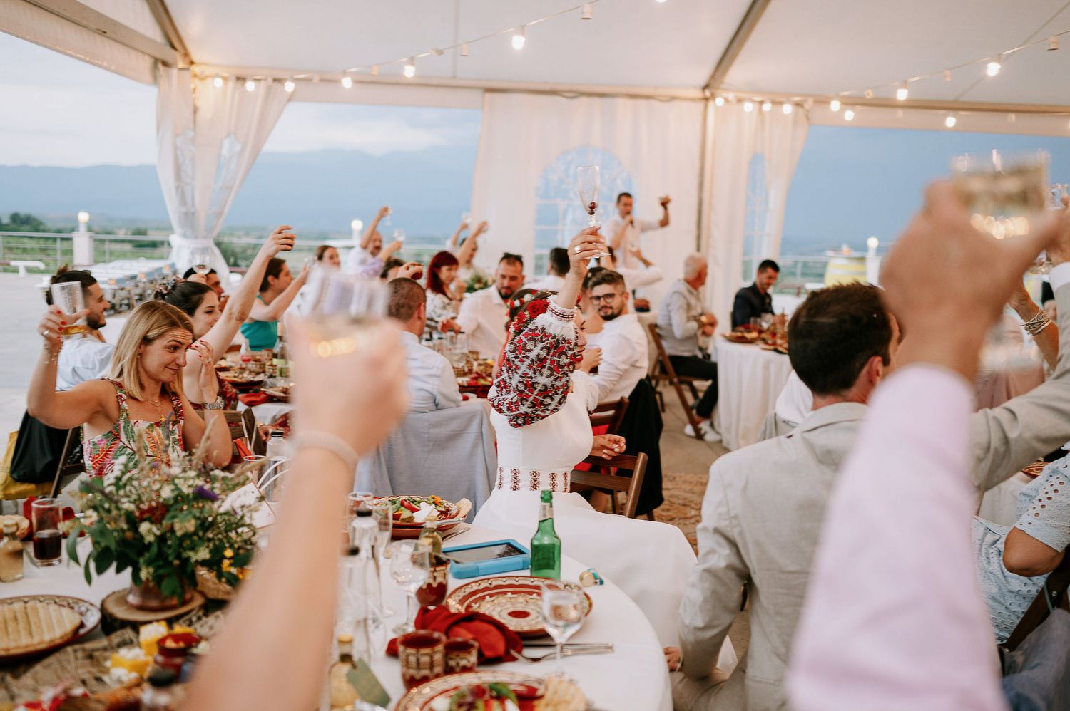 Enthusiastic wedding guests raising their hands during a toast at an outdoor tented reception.