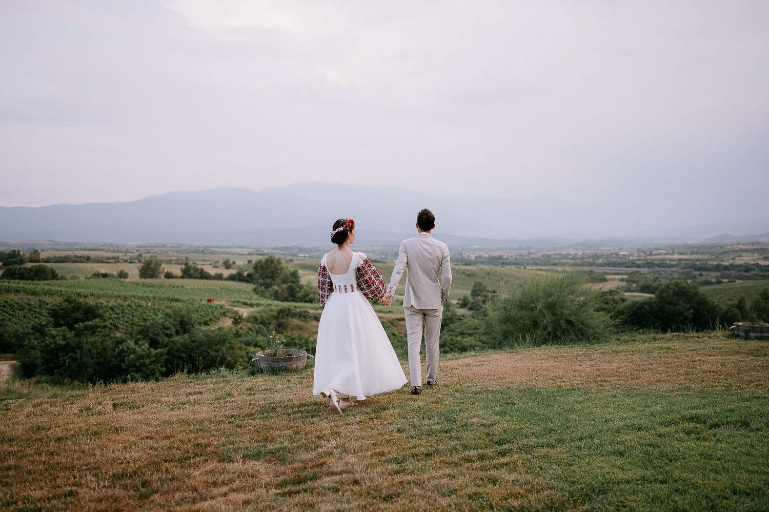 A couple in traditional wedding attire walks along a scenic hilltop overlooking a valley landscape.