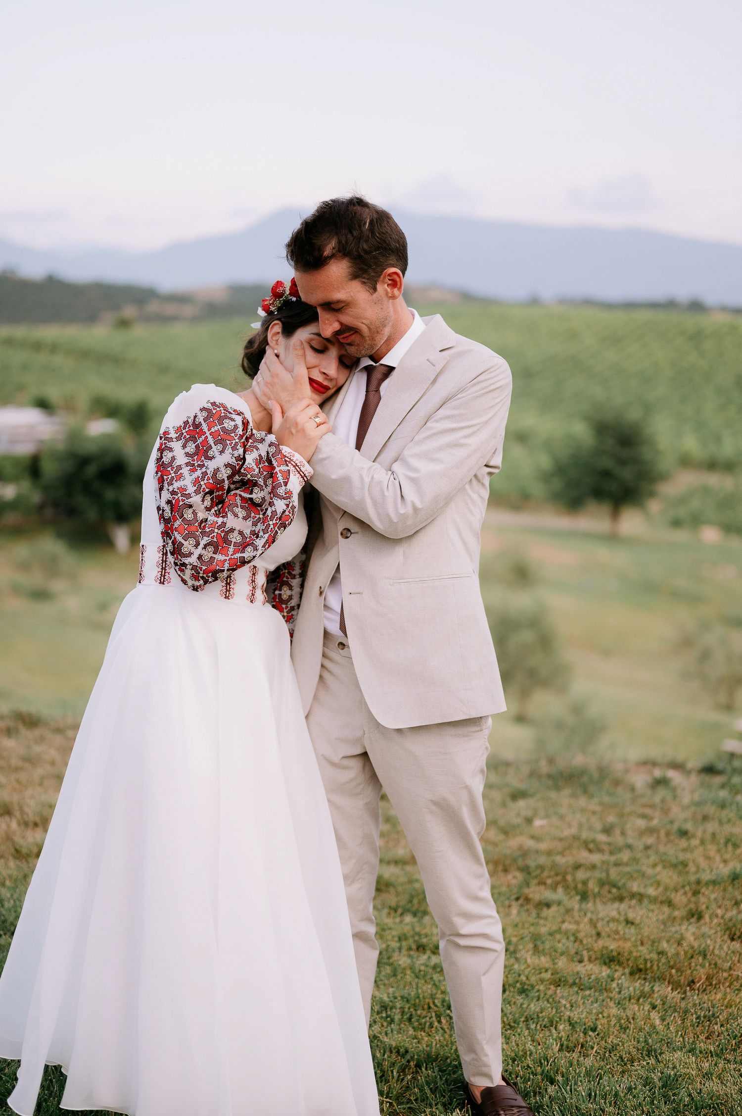 A tender wedding moment captured in a green field with mountains in the background.