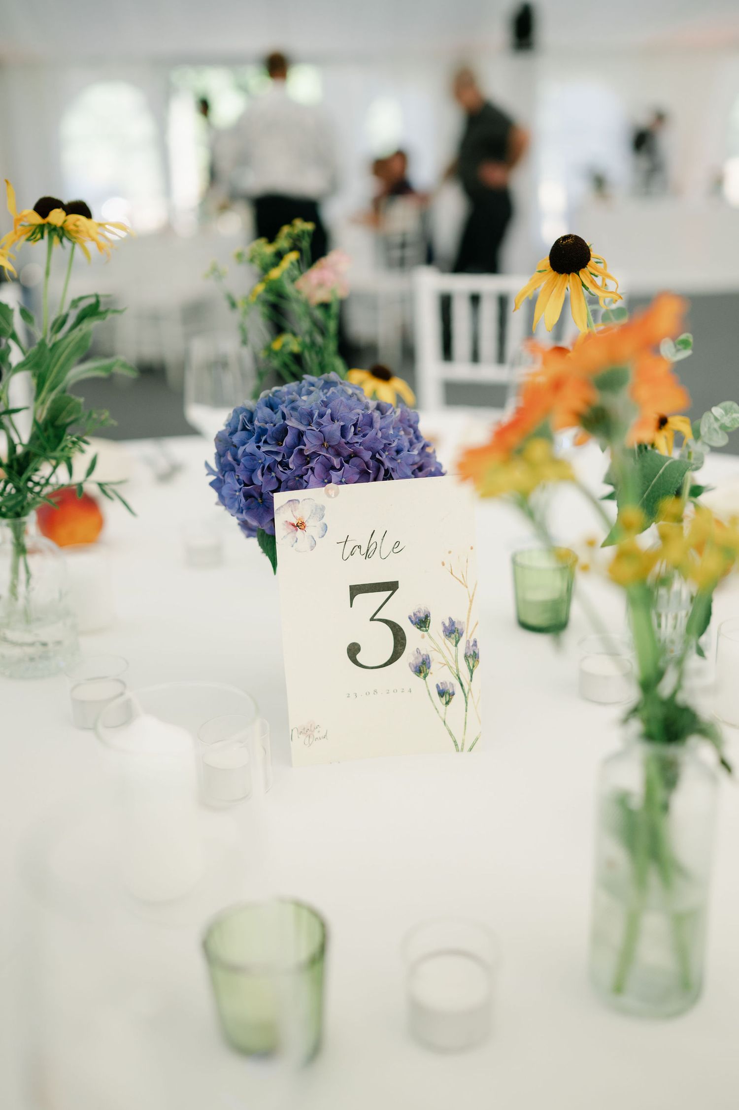 White table settings with colorful wildflowers in glass vases and table number cards at an elegant wedding reception.
