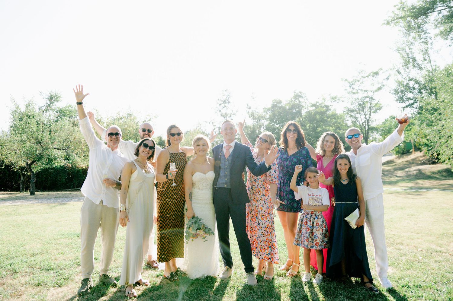 Large wedding group gathers for photos on a sunny hillside with trees in background.