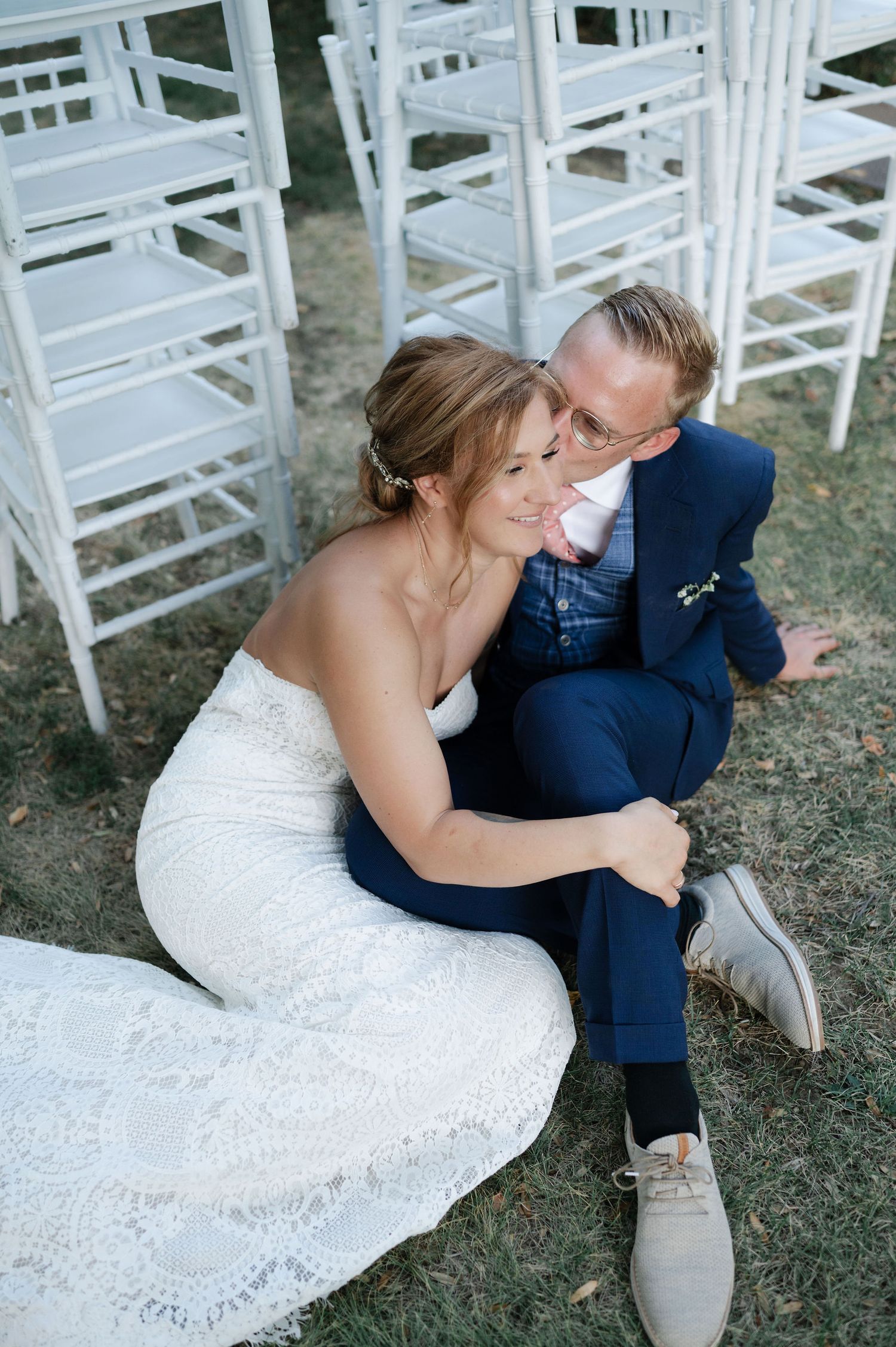 Wedding couple embraces on lawn with white ceremony chairs in background.