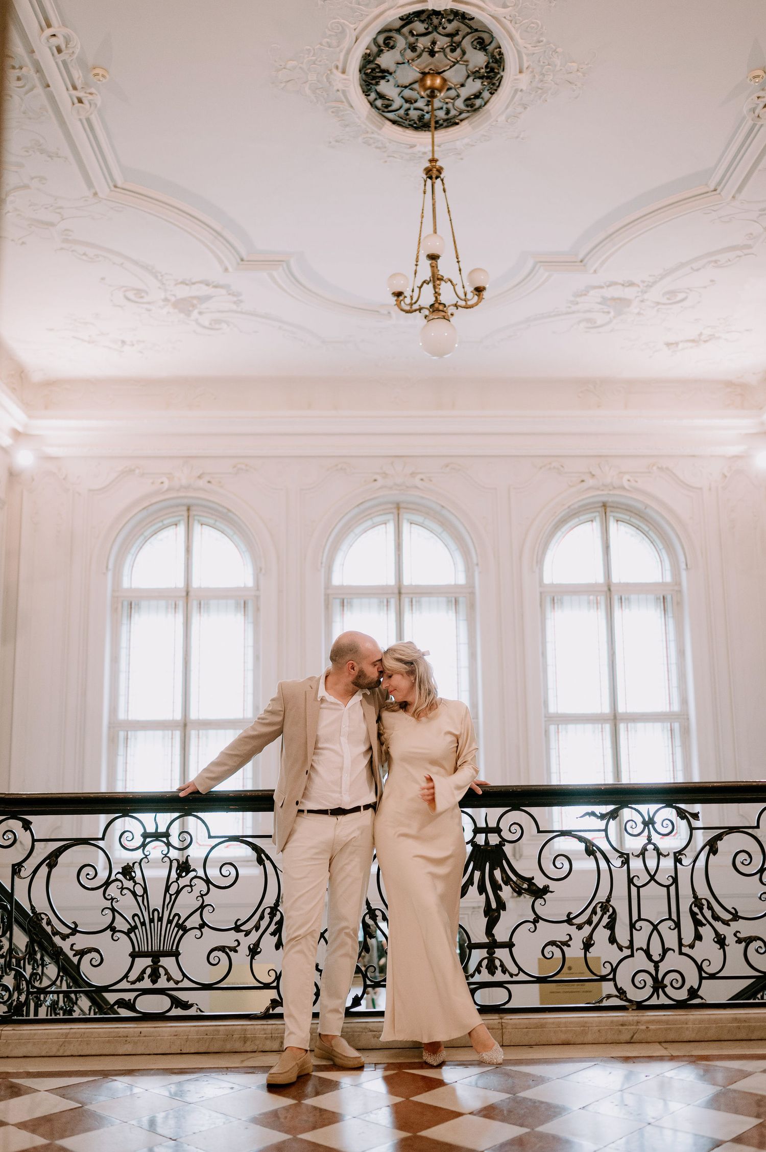 A couple shares an intimate moment in a luxurious white room with chandelier and checkerboard floor.